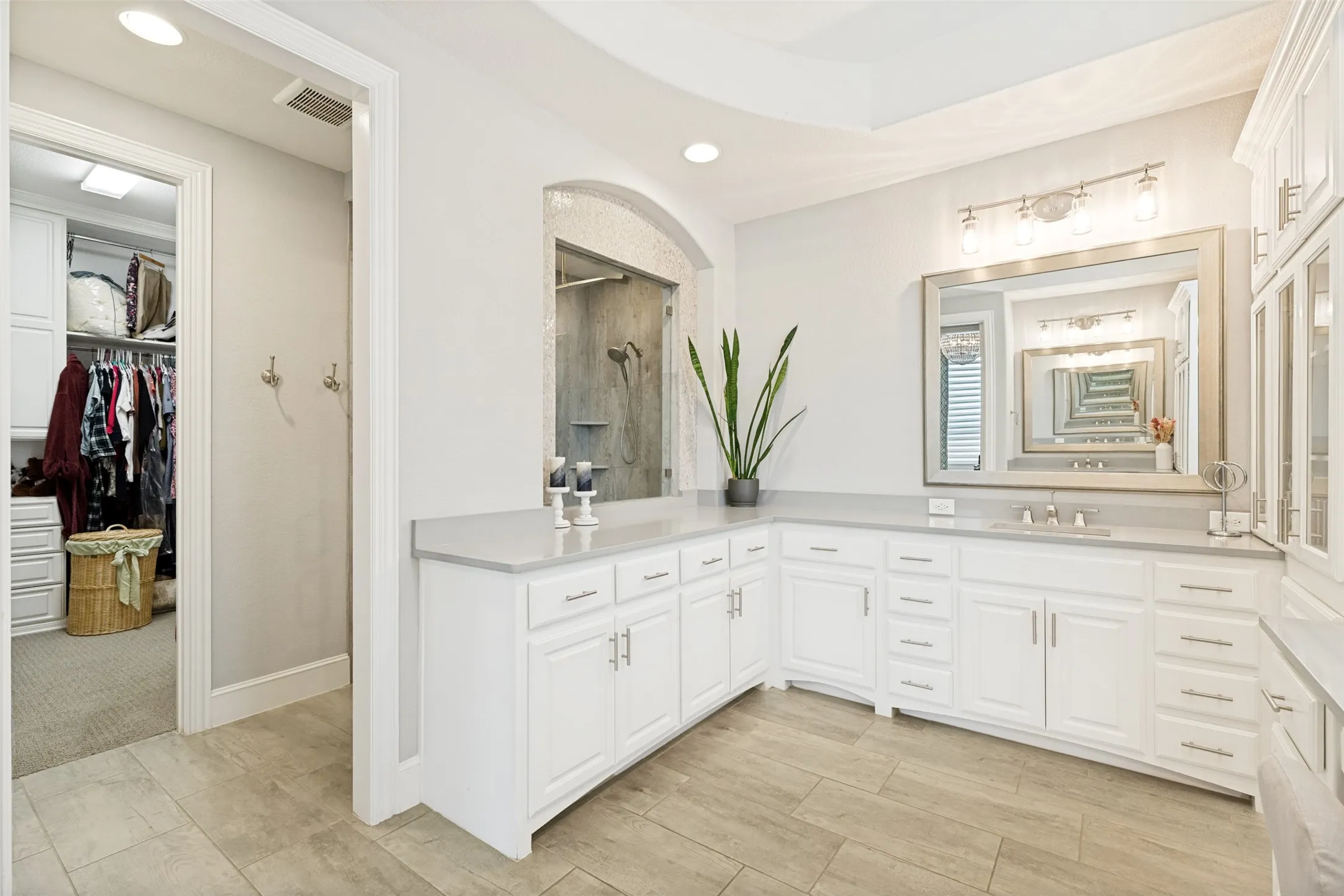 The cabinets wrap around the room to add a third vanity space with a sink and mirror as well as extra storage.  A window is open to the shower to provide light.  The primary closet has custom cabinetry including cabinets, drawers and shelving, and the closet opens to the hallway with the utility room.