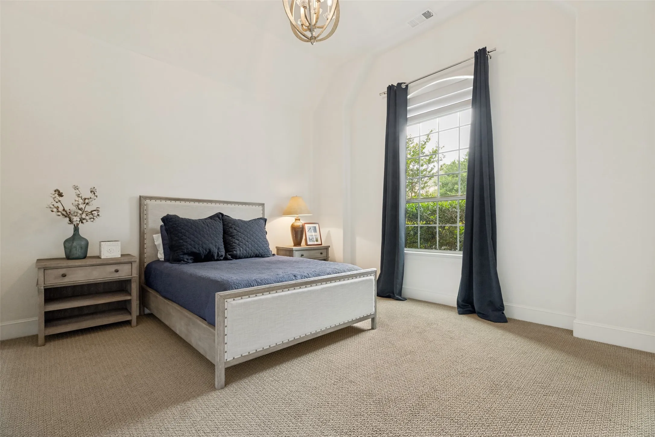 The second downstairs bedroom is ideal for guests, a nursery on the same floor as the primary suite or a teen sanctuary. Berber-style carpet is on the floor and a pretty hanging light fixture adds an elegant touch.  An arched window to the front of the home has drapery panels and shades.  Not seen in the photo is a built-in bookcase secluded by the entrance door.
