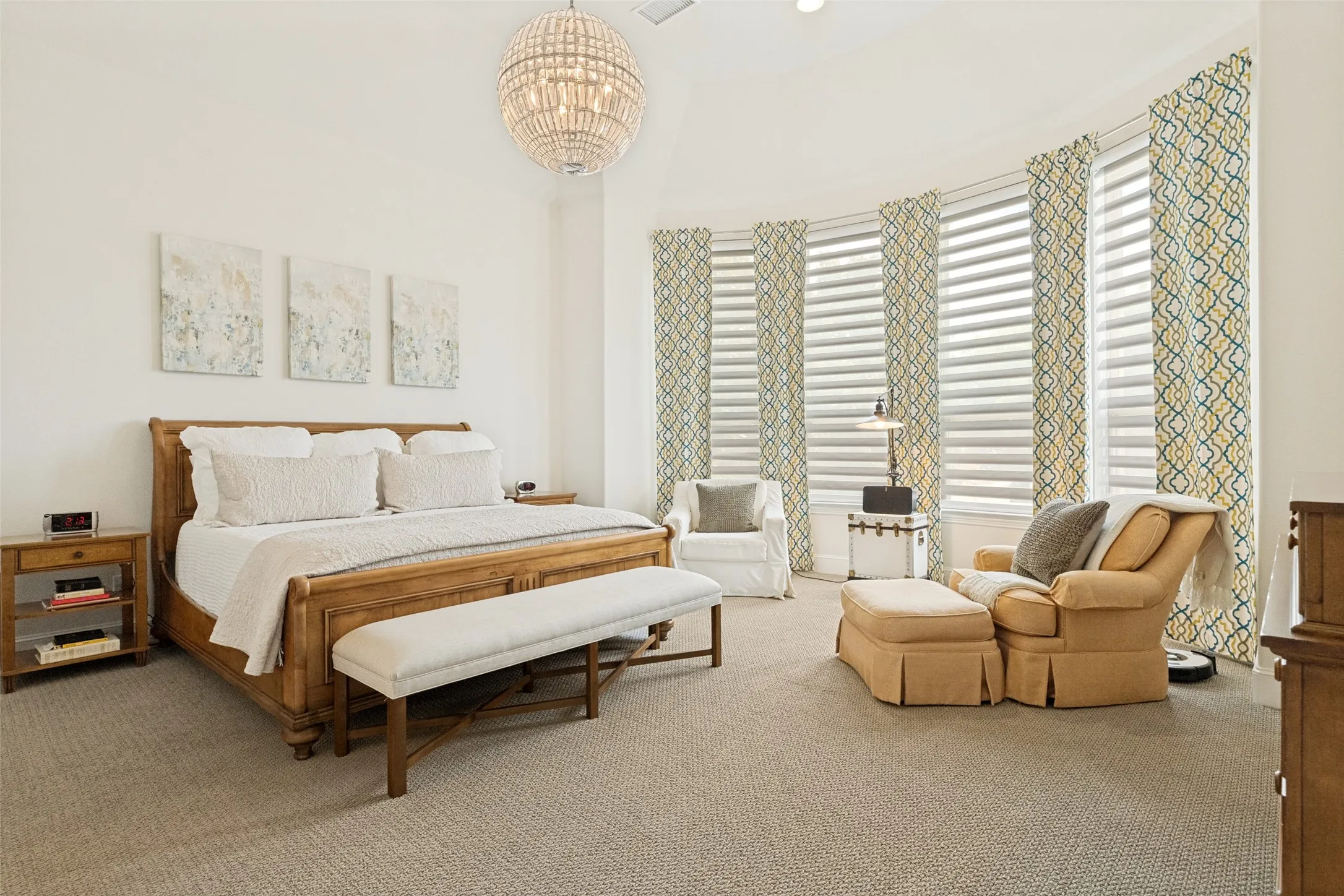 The primary bedroom is a haven of relaxation for homeowners.  An curved extended space with four windows allows room for a sitting area.  The windows have drapery panels and shades for privacy.  An elegant light fixture hangs from the raised ceiling and carpet is on the floor.  There is plenty of space for furniture in this primary bedroom.