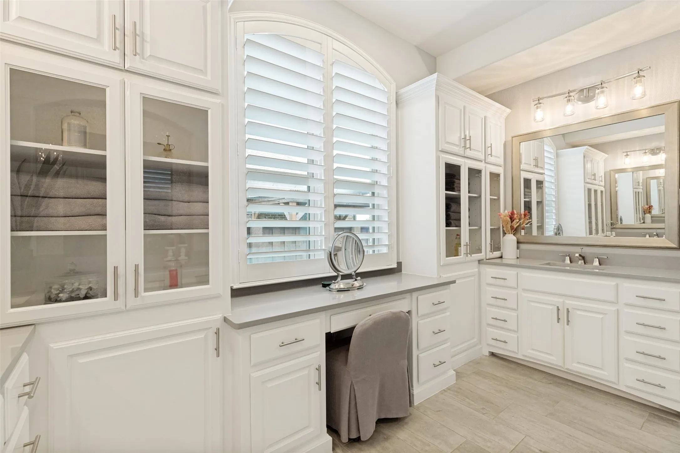 The primary suite includes a luxurious spa-inspired bath.  Abundant storage is available in cabinets that include towers with glass fronts and a third tier.  The white painted vanity has taupe countertops with a wide metallic-framed mirror above.  Light fixtures light the vanity.  A second vanity area includes a space for sitting with an arched window with shutters above.