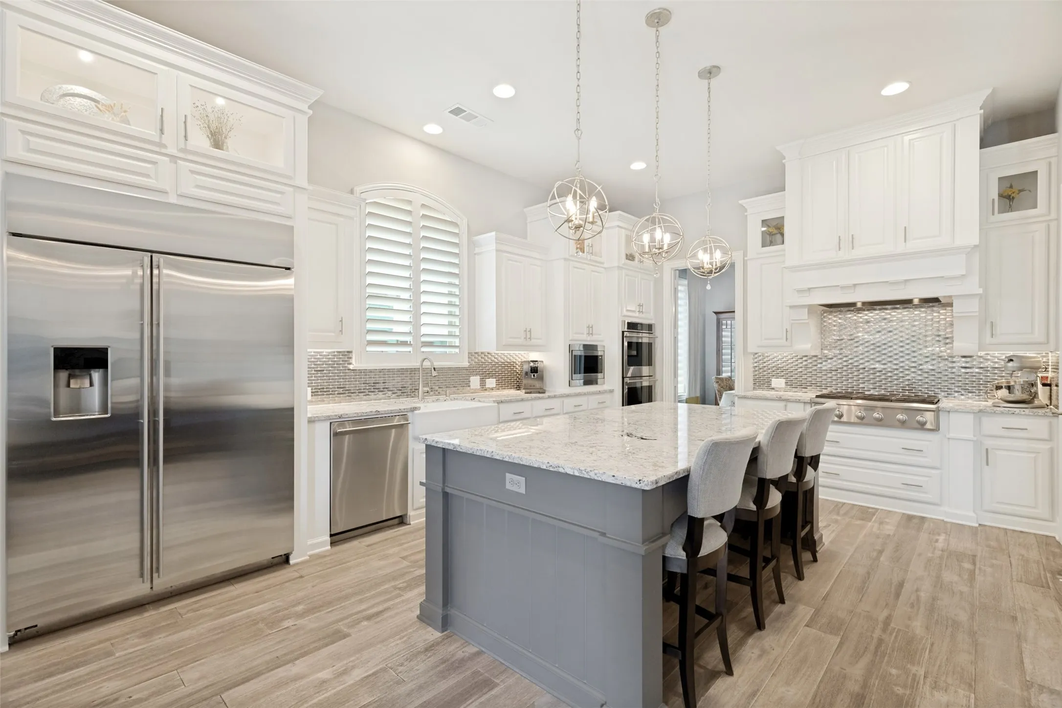 Abundant white cabinets soar almost to the ceiling with a third tier of cabinets with glass front and lights.  A striking vent hood rises above the six-burner commercial-style gas cooktop.  Metallic tiles in a basketweave design serve as backsplashes.  A trio of attractive pendant lights are above the island.