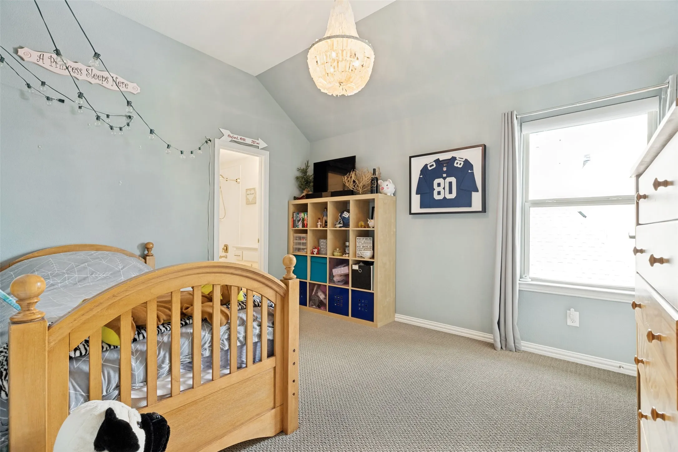 This bedroom views the side of the home.  It also have blue wall paint.  A ceiling light fixture adds an elegant touch.  The window has shades with drapery panels.  The third upstairs bedroom has a window with shutters.  It also has a ceiling fan with a light and it views the side of the home.