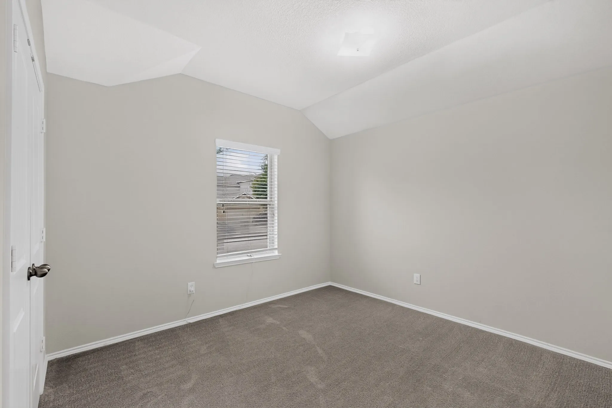 Neutral-toned walls, vaulted ceiling, single window with blinds, carpeted flooring, and white interior door with dark hardware