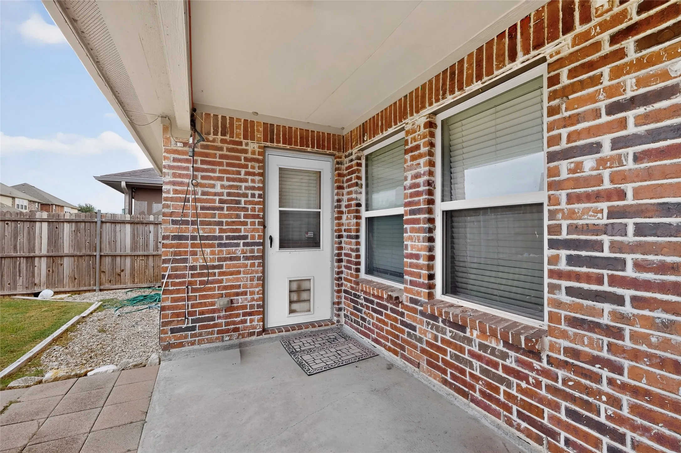 Covered concrete patio with a brick exterior, two windows featuring blinds, and a white door with a pet access panel