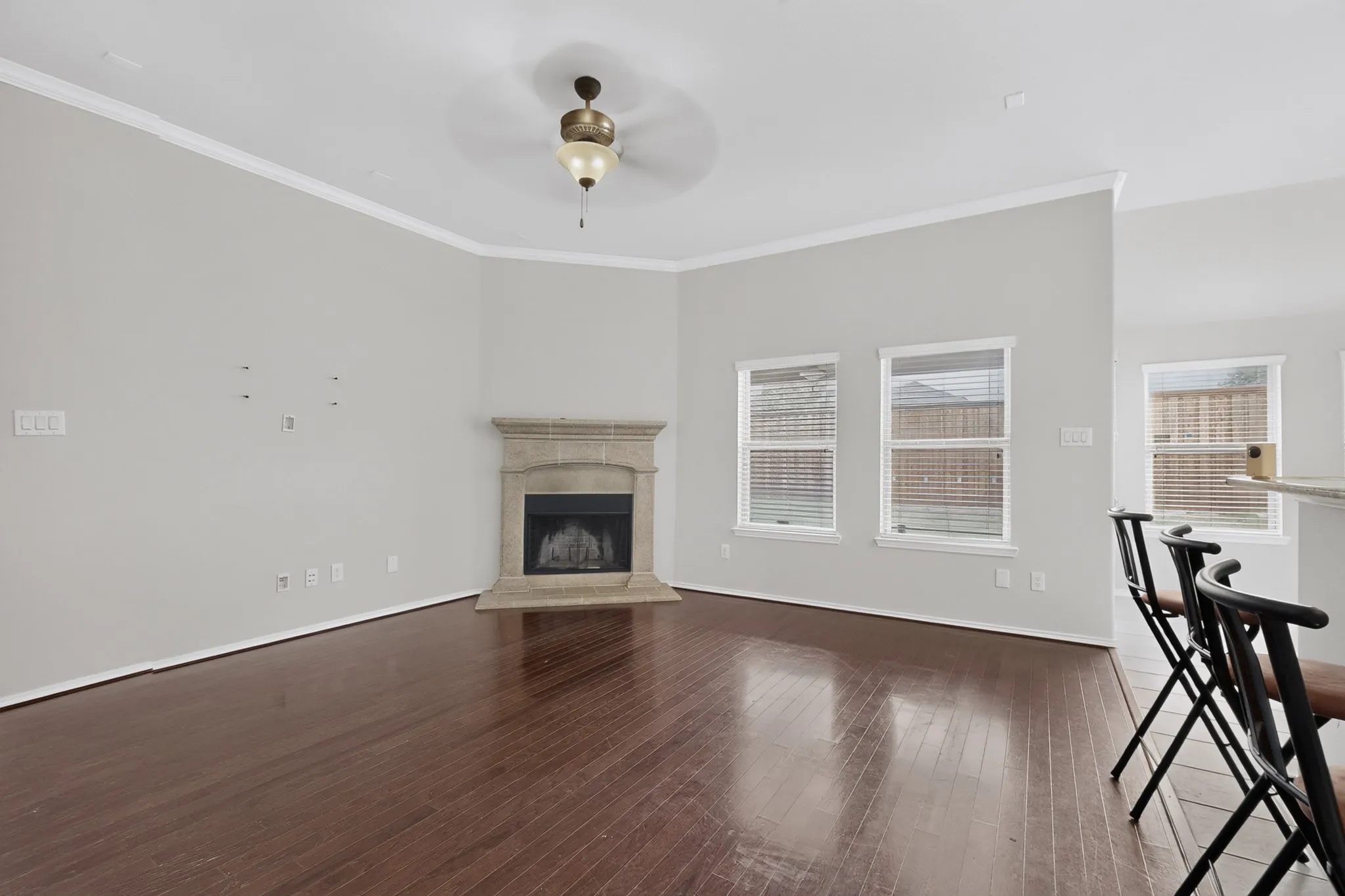 Wood-finish flooring, corner fireplace with stone surround, crown molding, ceiling fan, and multiple windows with blinds