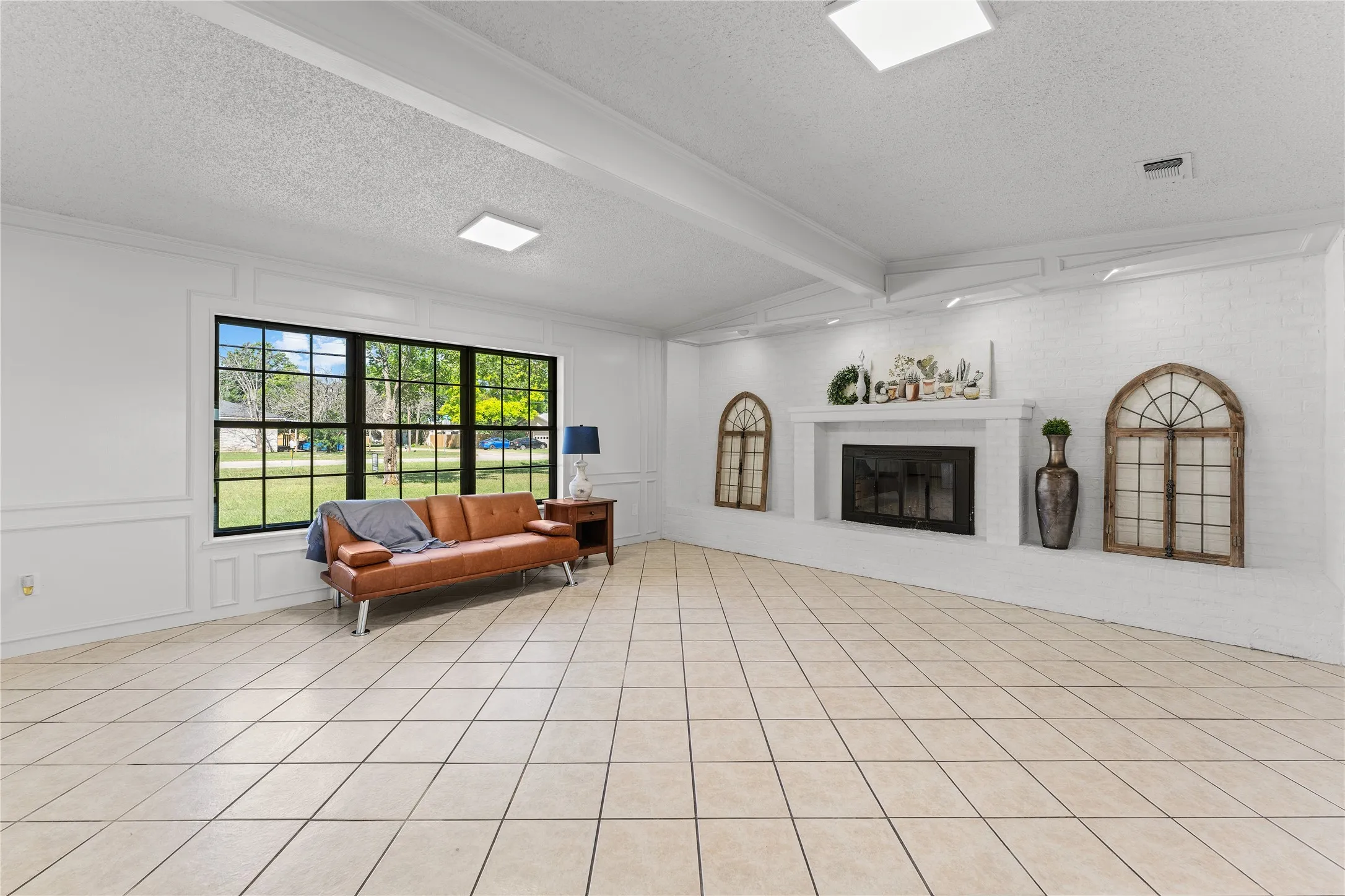 Spacious interior with neutral tile flooring, a large multi-pane window, and a white brick fireplace with a mantel