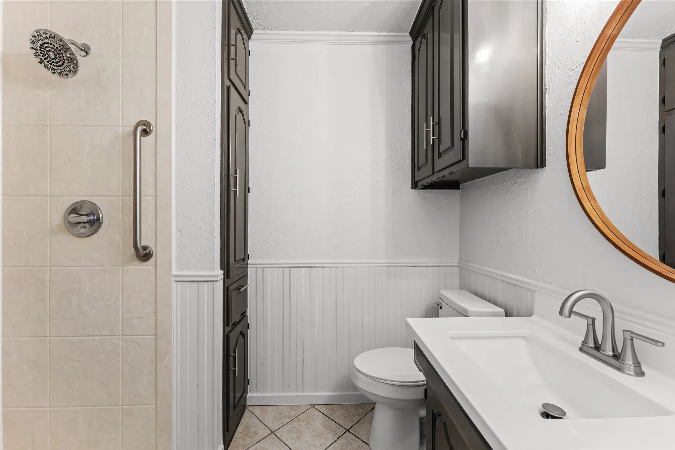 Bathroom featuring a tiled shower with grab bar and rain shower head, dark wood-finish cabinetry, a single vanity with an integrated sink, and white beadboard wainscoting