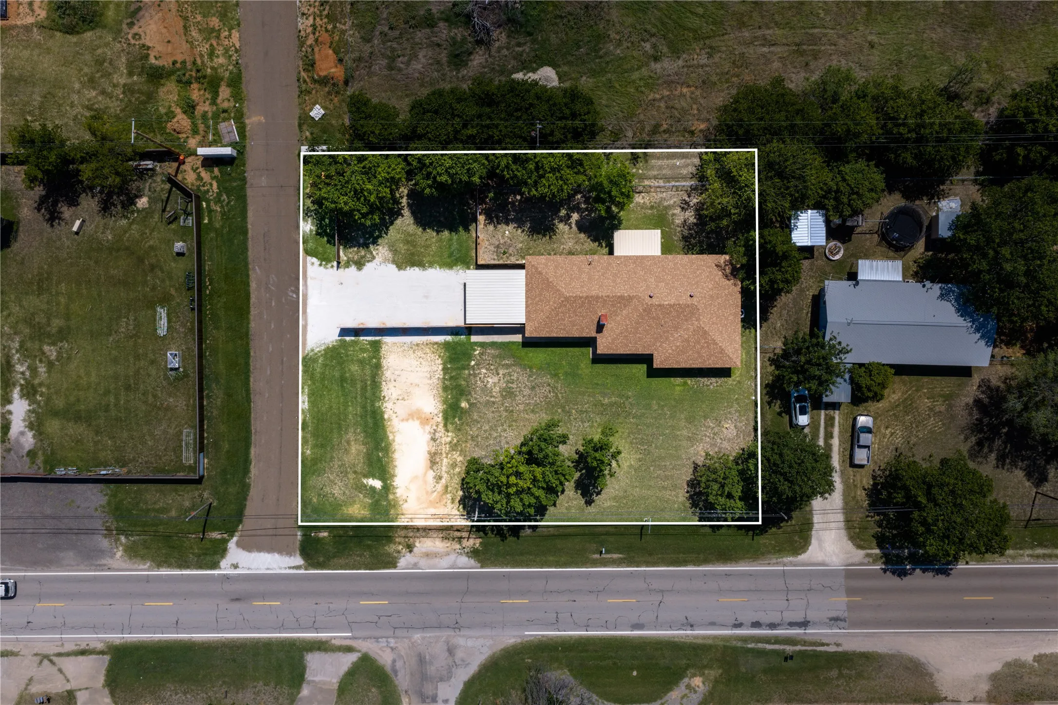 Aerial perspective of a property featuring a single-story residence with a brown shingle roof, set on a green lawn with scattered mature trees