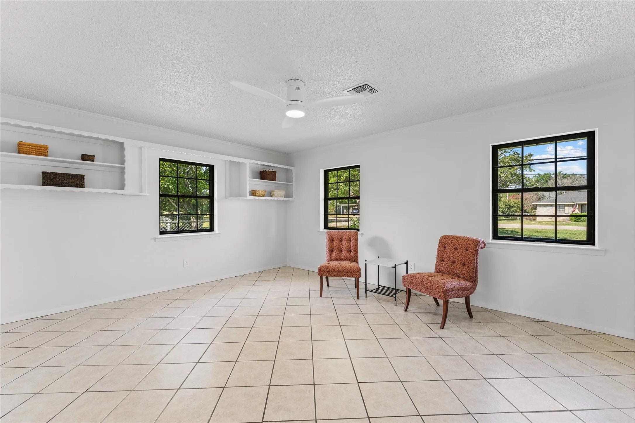 Spacious interior room featuring light-toned tile flooring, multiple windows with dark frames, and a ceiling fan
