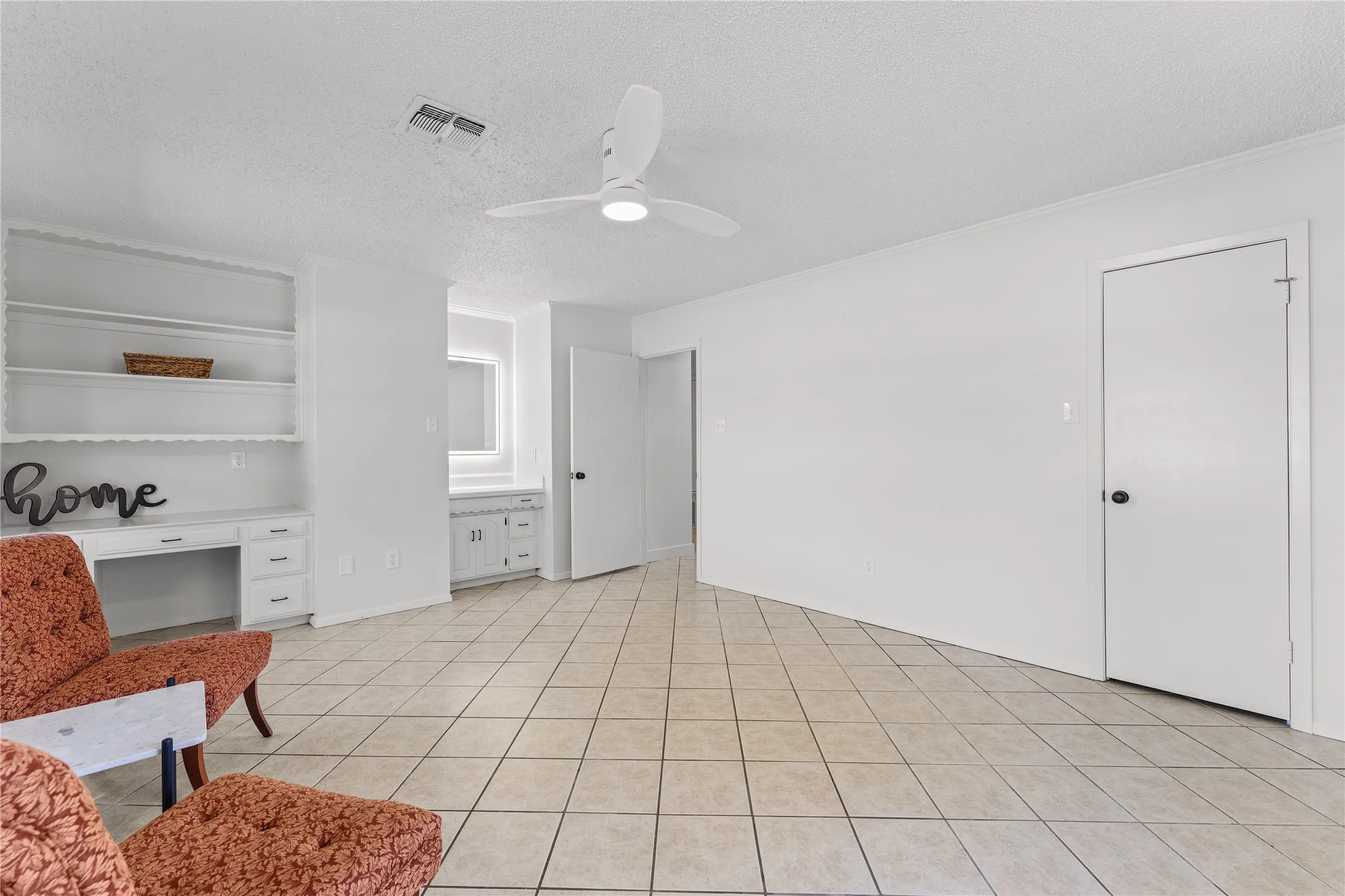 Spacious room featuring neutral tile flooring, built-in shelving with desk, and a ceiling fan
