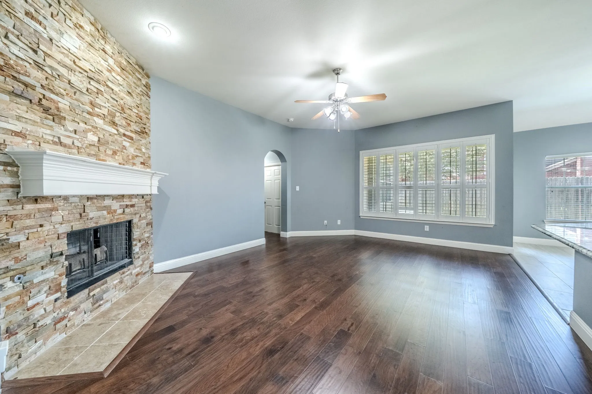 Spacious living area featuring a stacked stone fireplace with a white mantel, rich wood-finish flooring, and a ceiling fan with light