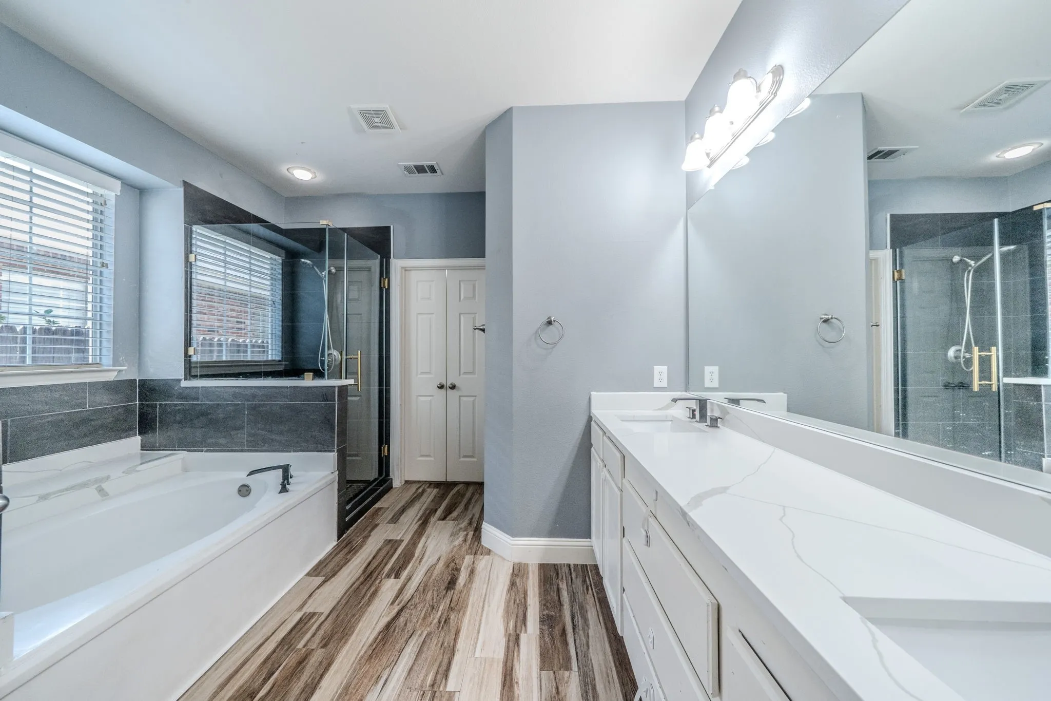 Bathroom featuring a dual vanity with a white countertop, a built-in tub, a glass-enclosed shower, wood-finish flooring, and light blue walls