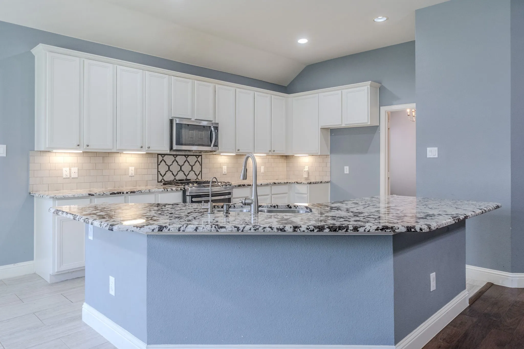Kitchen featuring a tiered island with a stainless steel sink and gooseneck faucet, white shaker cabinetry with under-cabinet lighting, subway tile backsplash, stainless steel microwave, and wood-finish flooring
