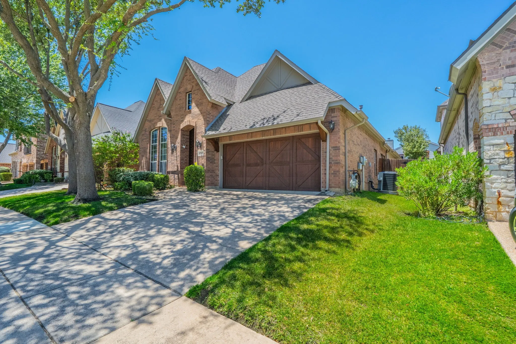 Brick and stone facade residence featuring a peaked roofline, a two-car garage with wood-finish doors, a concrete driveway, and mature landscaping