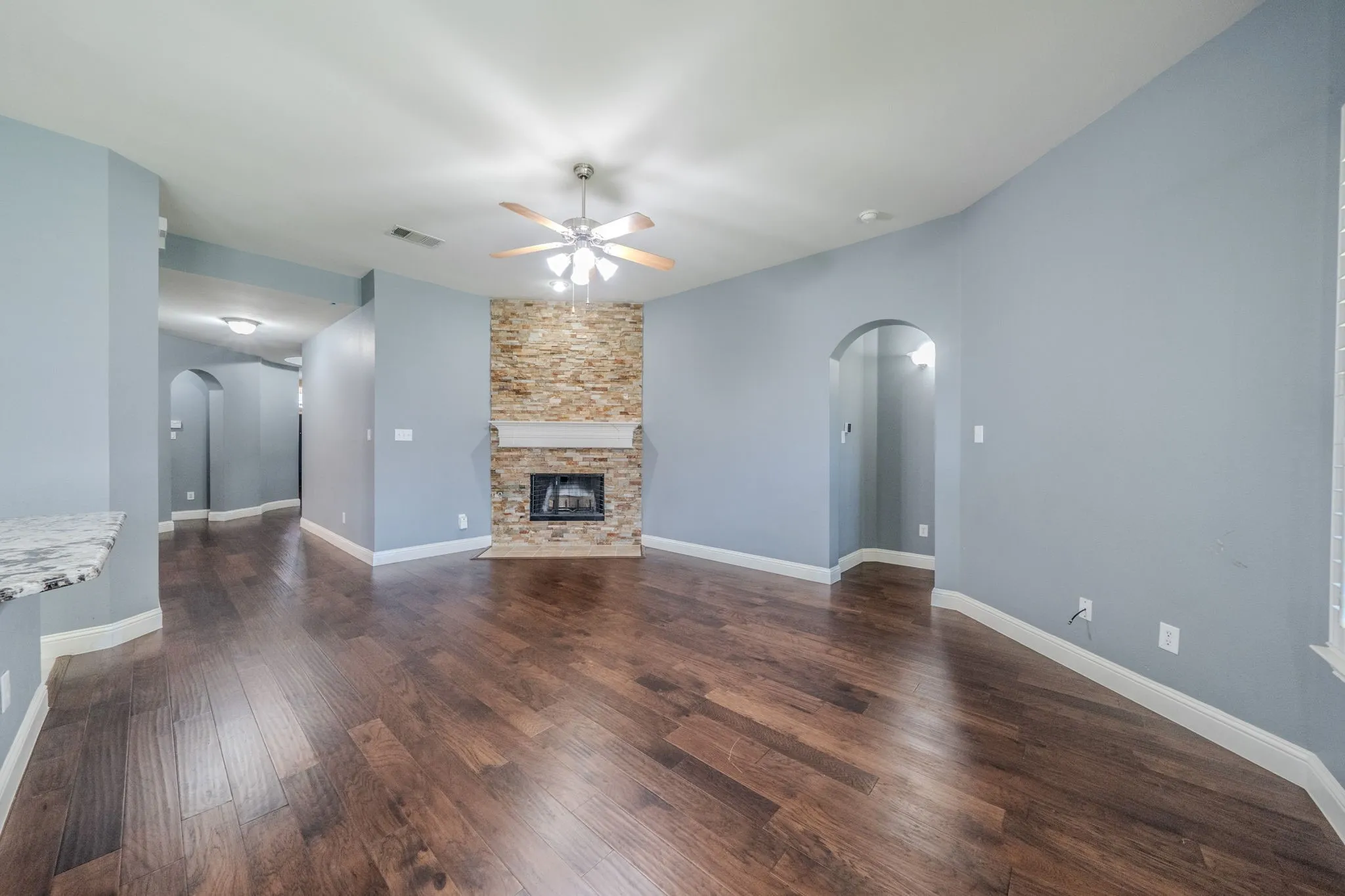 Spacious interior featuring wood-finish flooring, a stacked stone fireplace with a white mantel, and a ceiling fan with integrated lighting