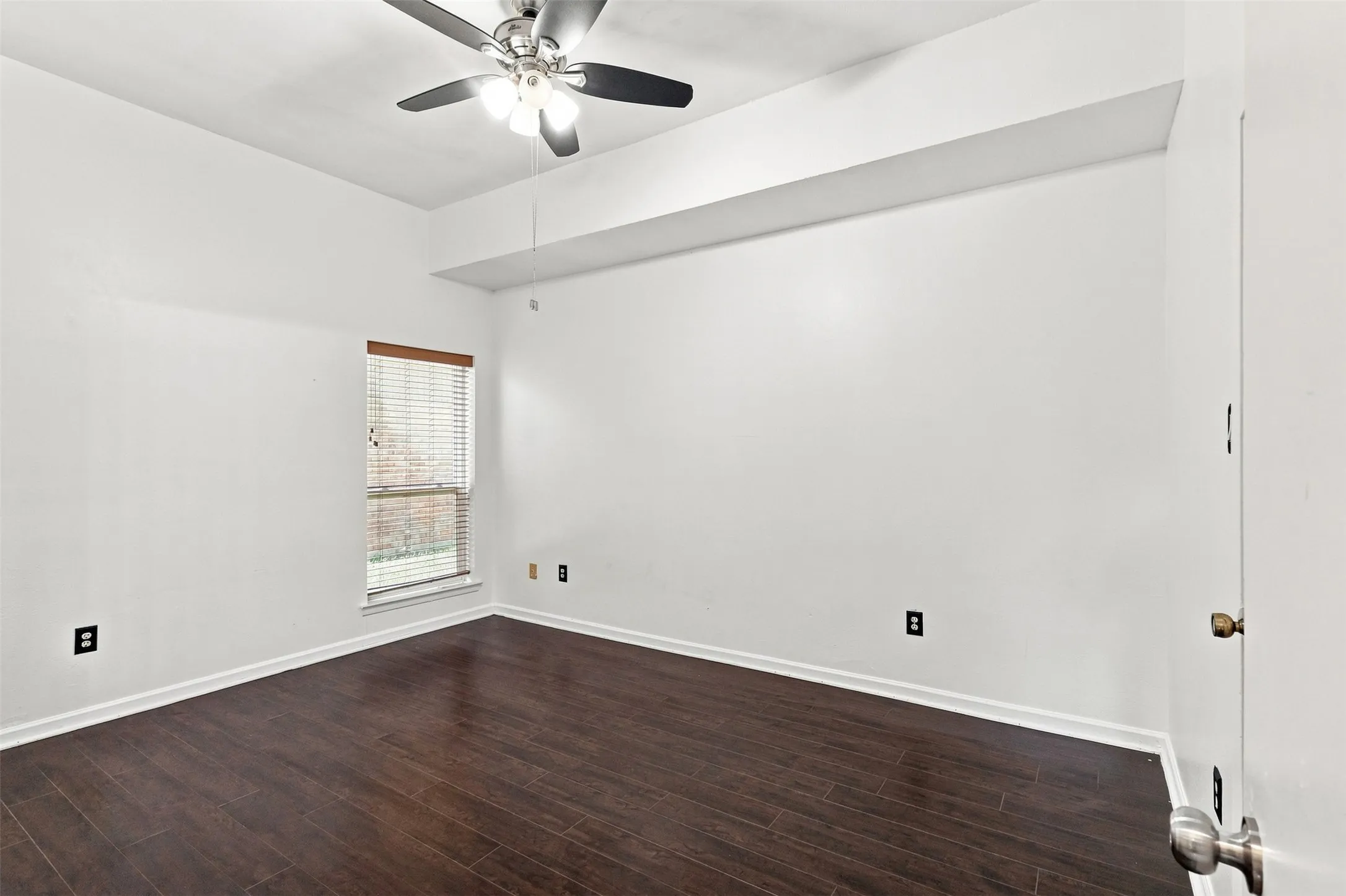 Room featuring dark wood-finish flooring, white walls, a window with blinds, and a ceiling fan with integrated lighting