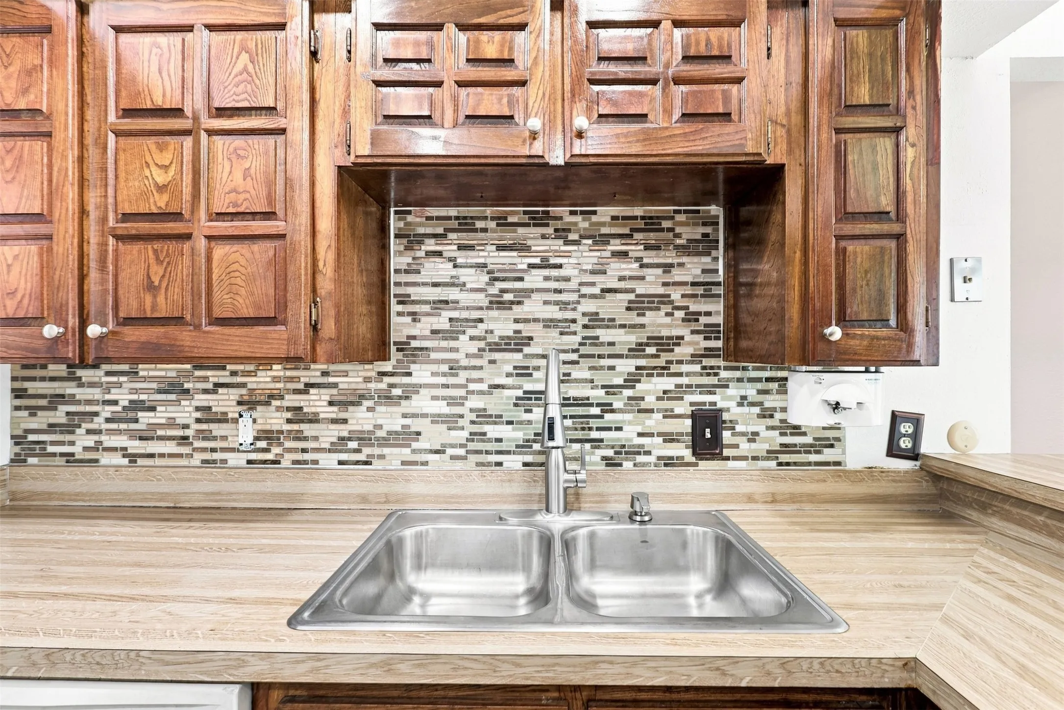 Wood-finish countertops, stainless steel double-basin sink, contemporary faucet, mosaic tile backsplash, and paneled wood cabinetry