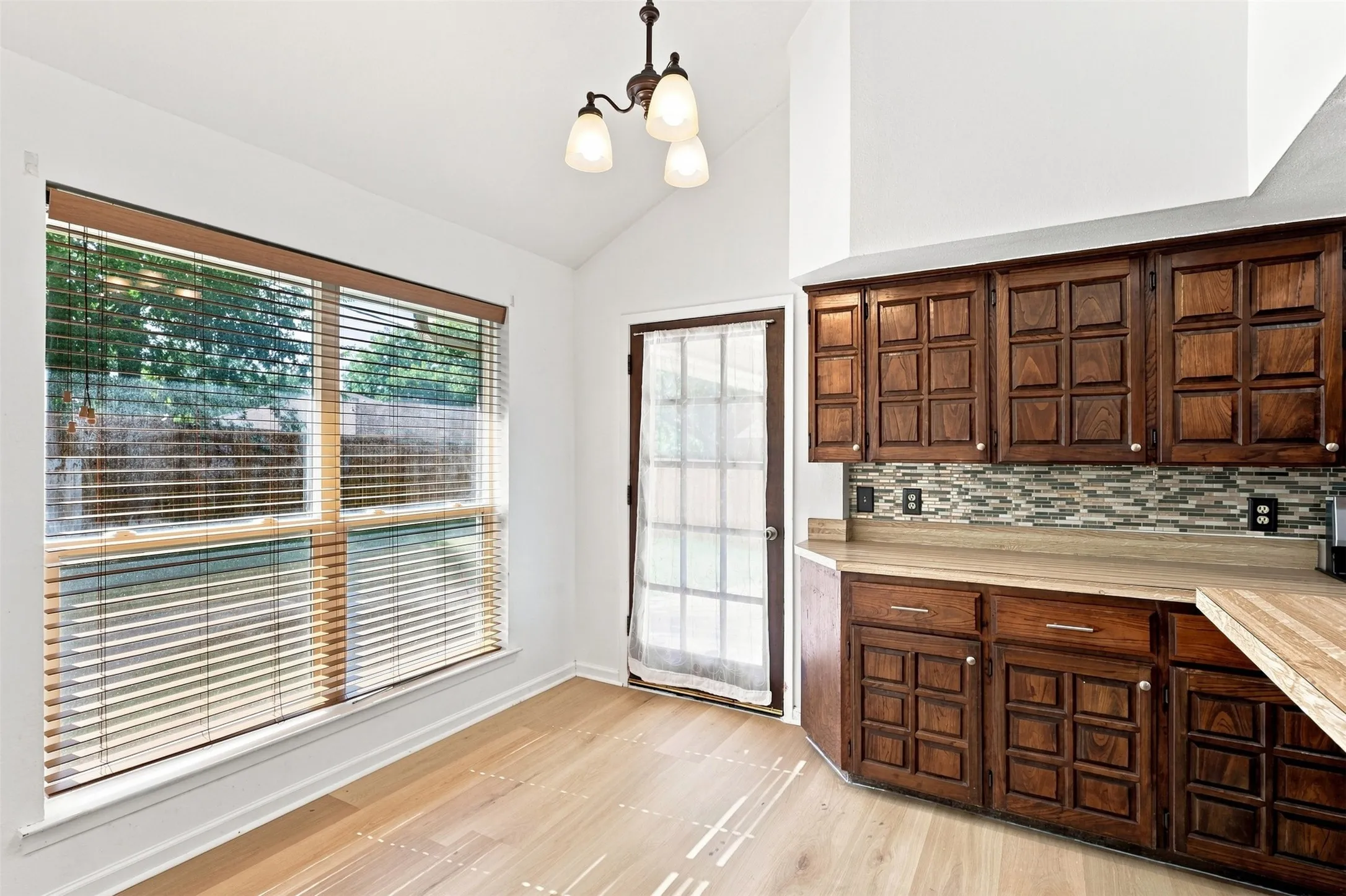 Vaulted ceiling, wood-finish flooring, wood cabinetry, multi-tone tile backsplash, and a glass-paneled door