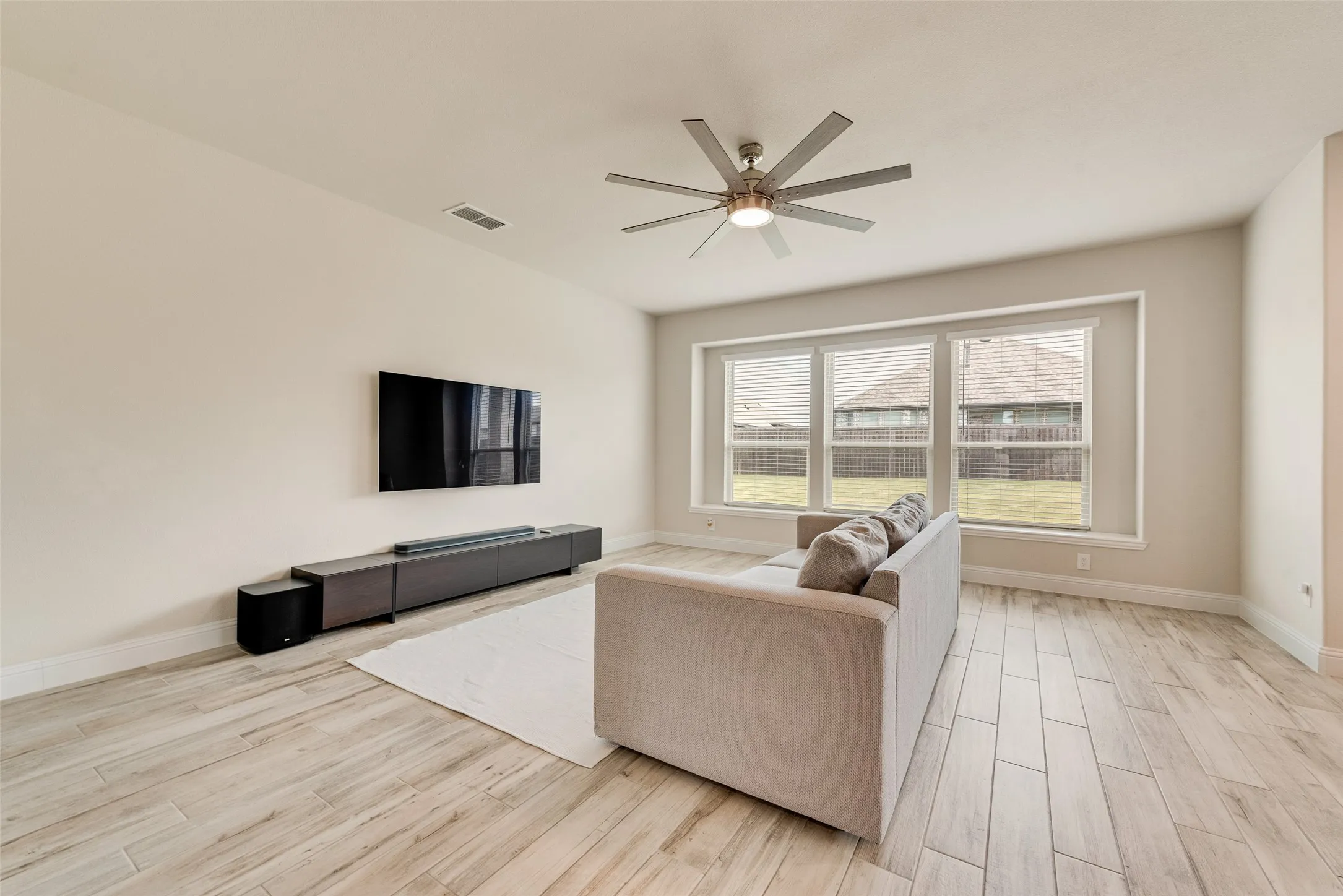 Living area featuring wood-look tile flooring, neutral wall tones, a contemporary ceiling fan, and a wall of three large windows providing abundant natural light