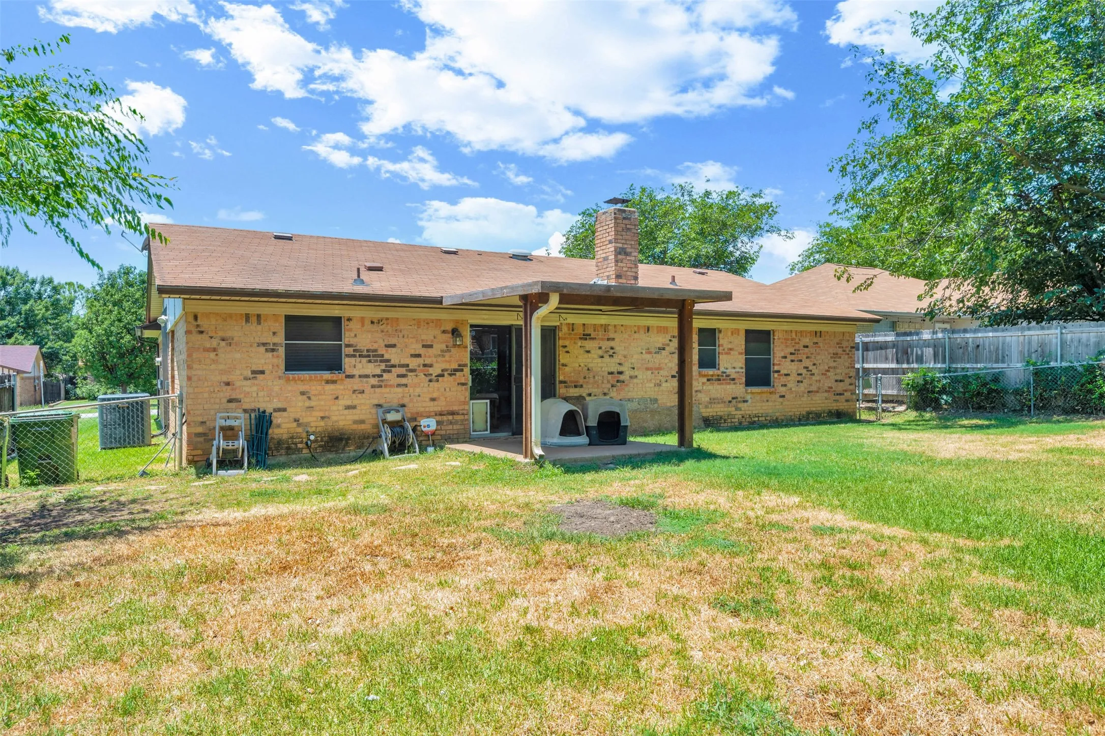 Expansive rear yard featuring a brick facade home with a covered patio, a prominent brick chimney, and a mix of manicured and natural grass areas