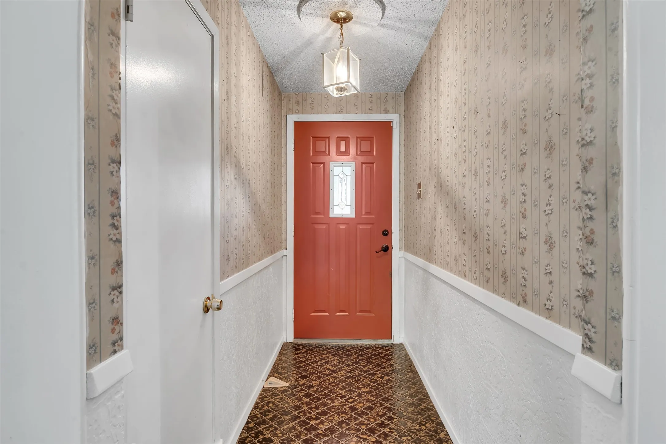 Entryway featuring a red paneled door with decorative glass inserts, patterned wallpaper, textured wainscoting, and a ceiling-mounted light fixture