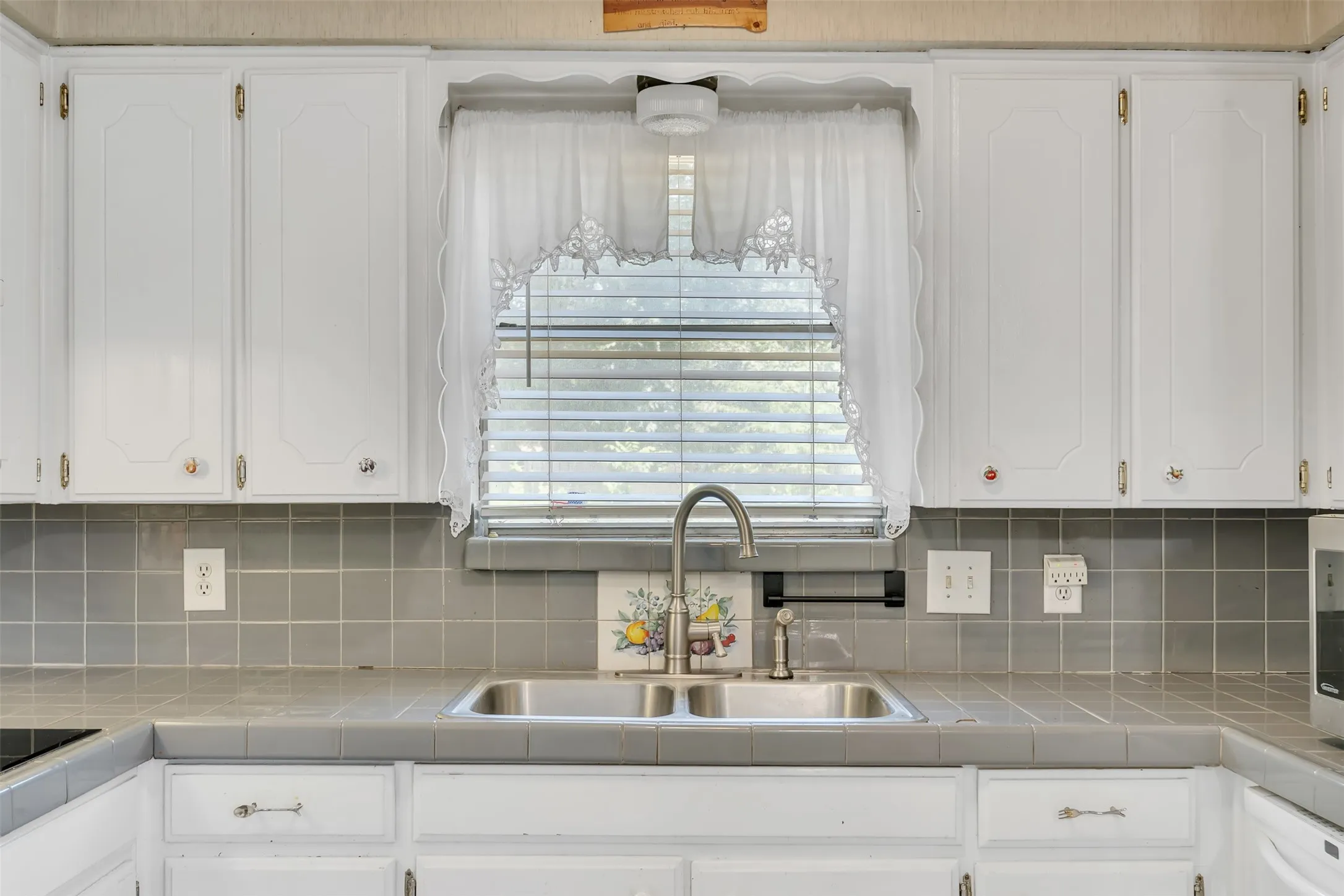 Kitchen sink area with a double basin stainless steel sink, gooseneck faucet, white cabinetry, and gray tiled countertops