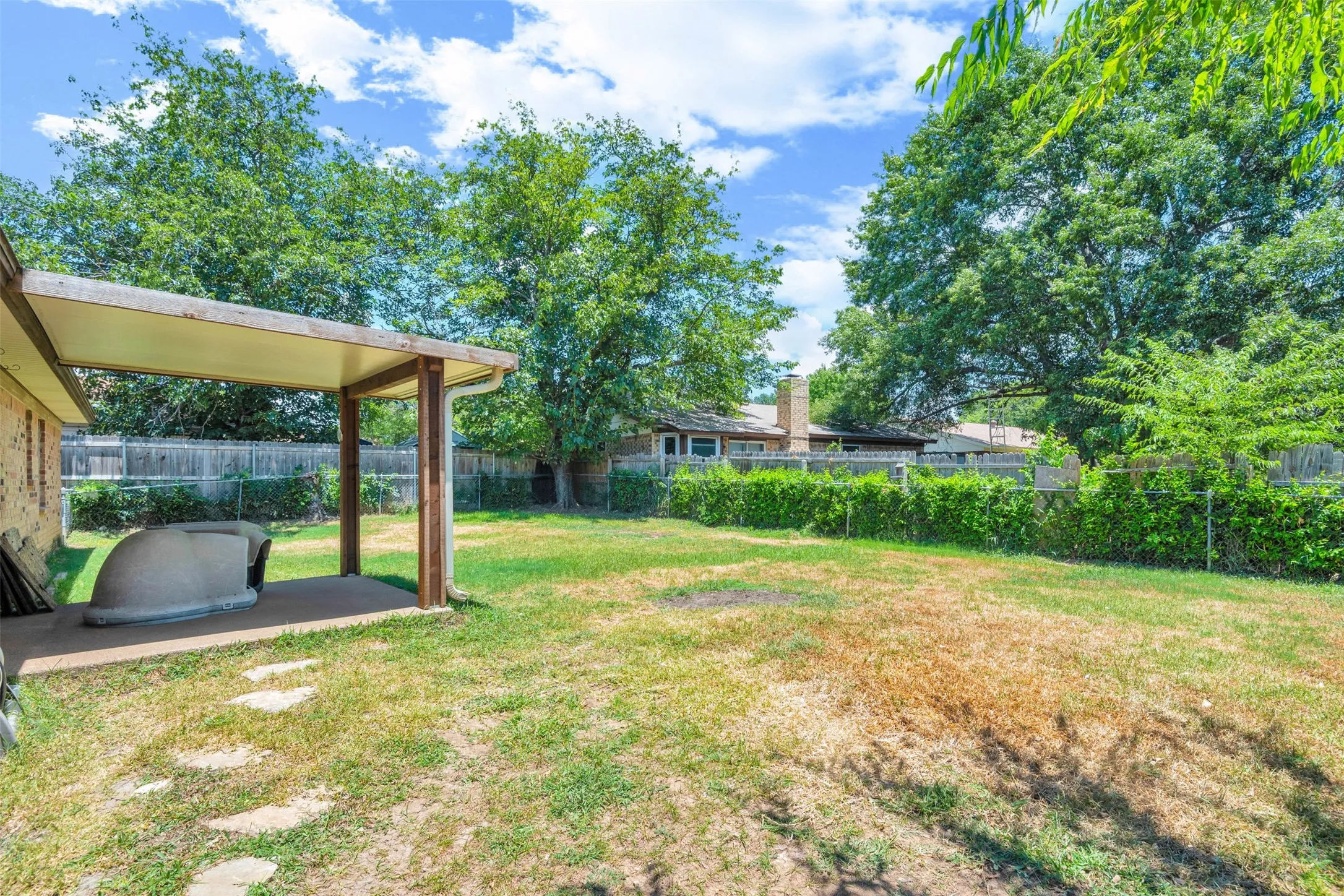 Expansive backyard featuring a covered patio with concrete flooring, stone pavers, mature trees, and a chain-link fence bordering lush green foliage