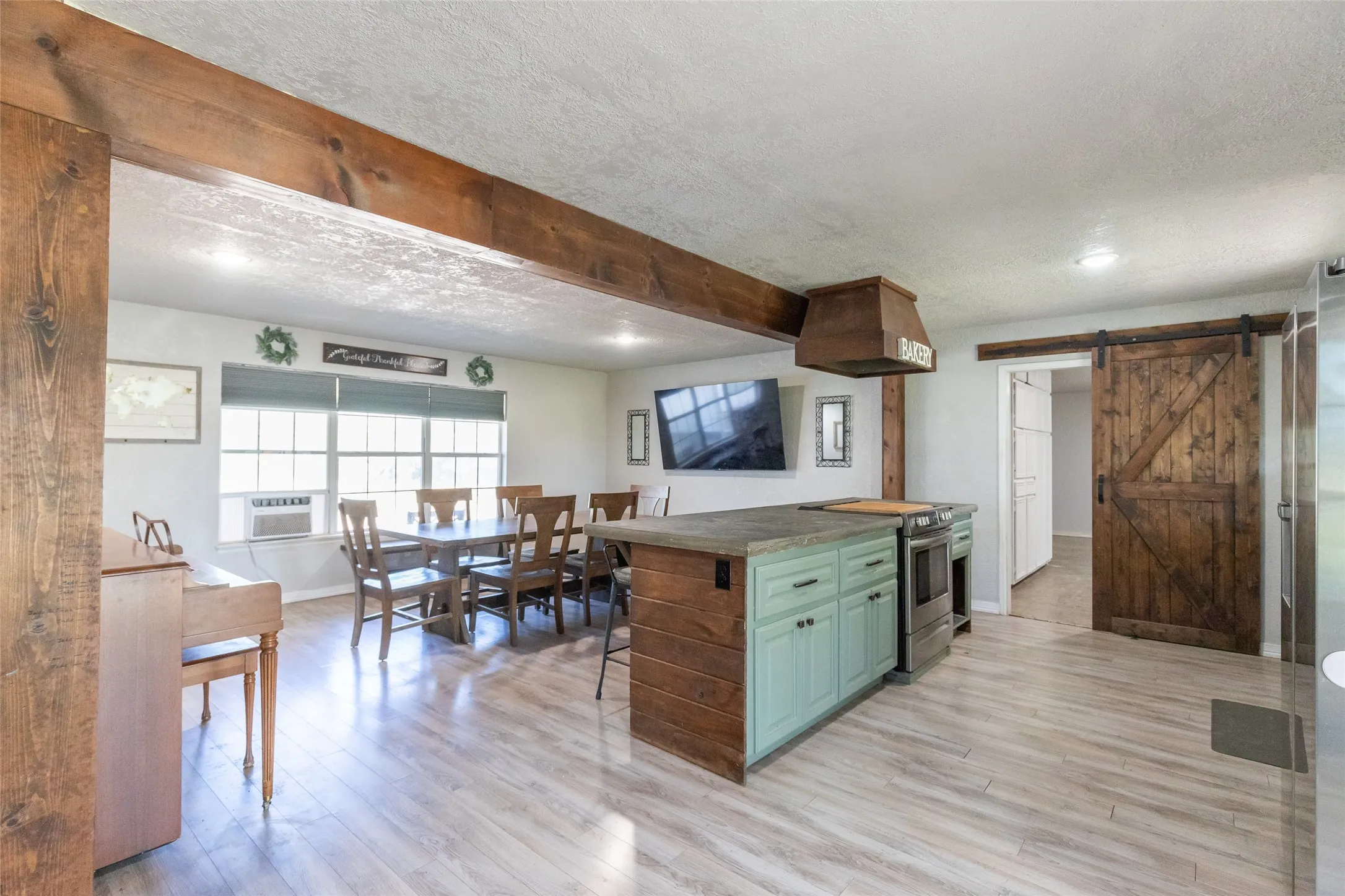 Open-concept kitchen and dining area featuring wood-finish flooring, exposed ceiling beams, a kitchen island with mint green cabinetry, and a sliding barn door