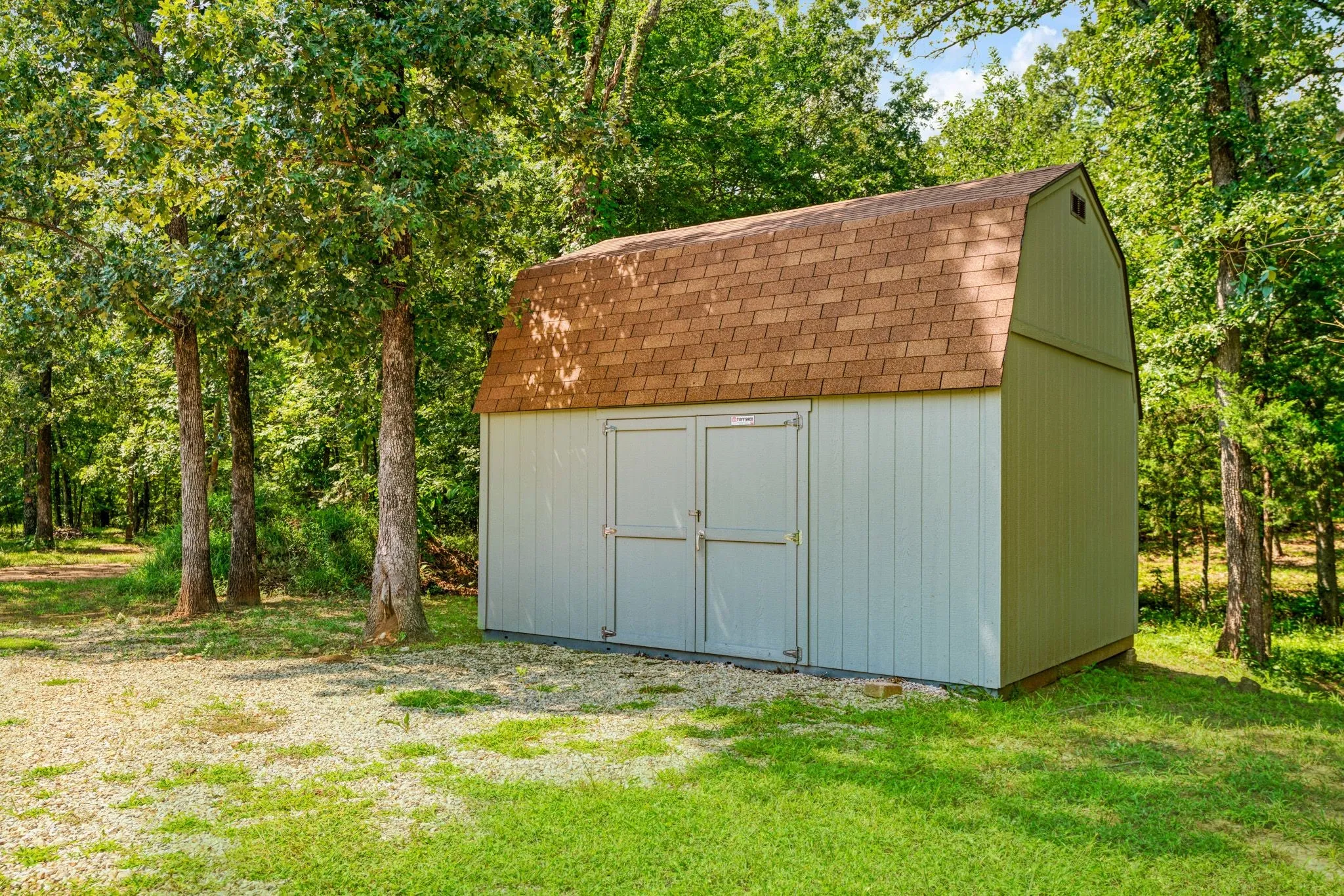 Gable roof storage shed featuring two front entry doors, vertical panel siding, and a surrounding lawn with mature trees