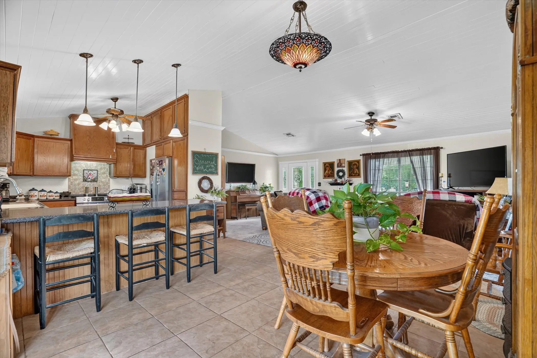 Open-concept kitchen and living area featuring tile flooring, wooden cabinetry, a breakfast bar with pendant lighting, and a beadboard ceiling