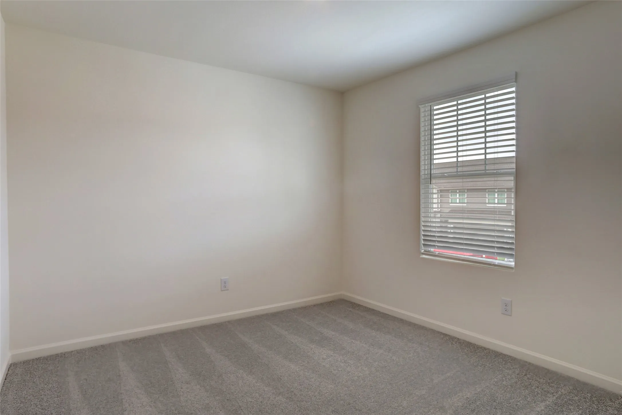 Light-filled room featuring neutral wall paint, gray patterned carpeting, a single window with blinds, and white baseboards
