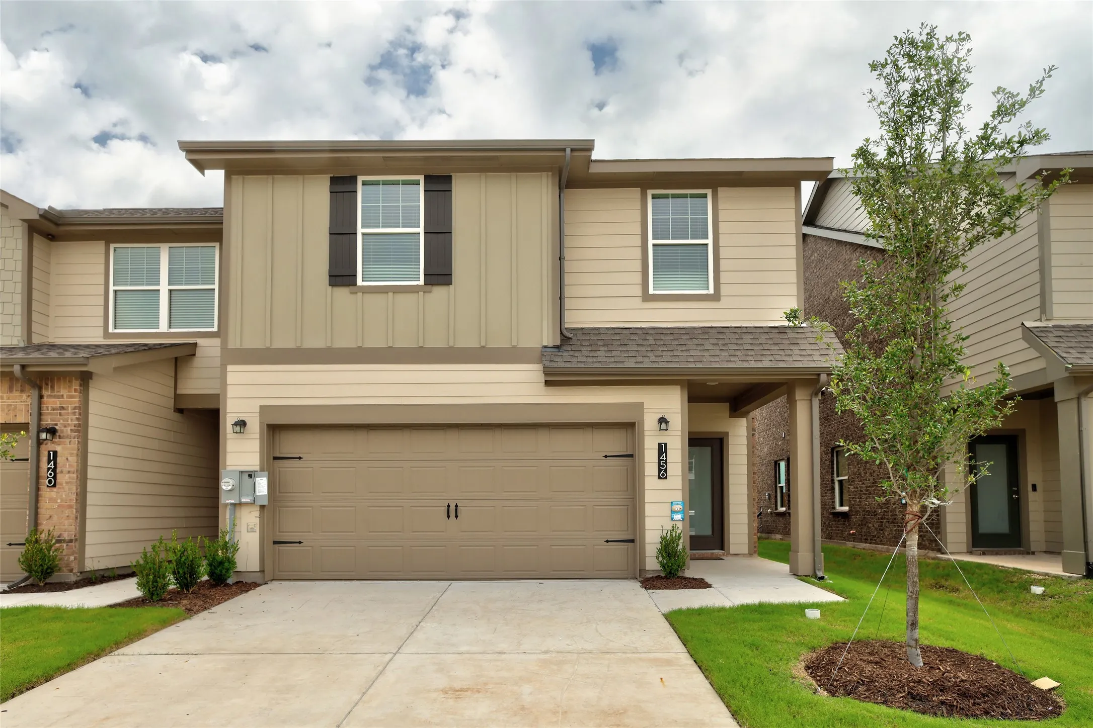 Contemporary two-story exterior featuring tan siding with vertical accents, dark shutters, an attached garage with a concrete driveway, and a covered entry