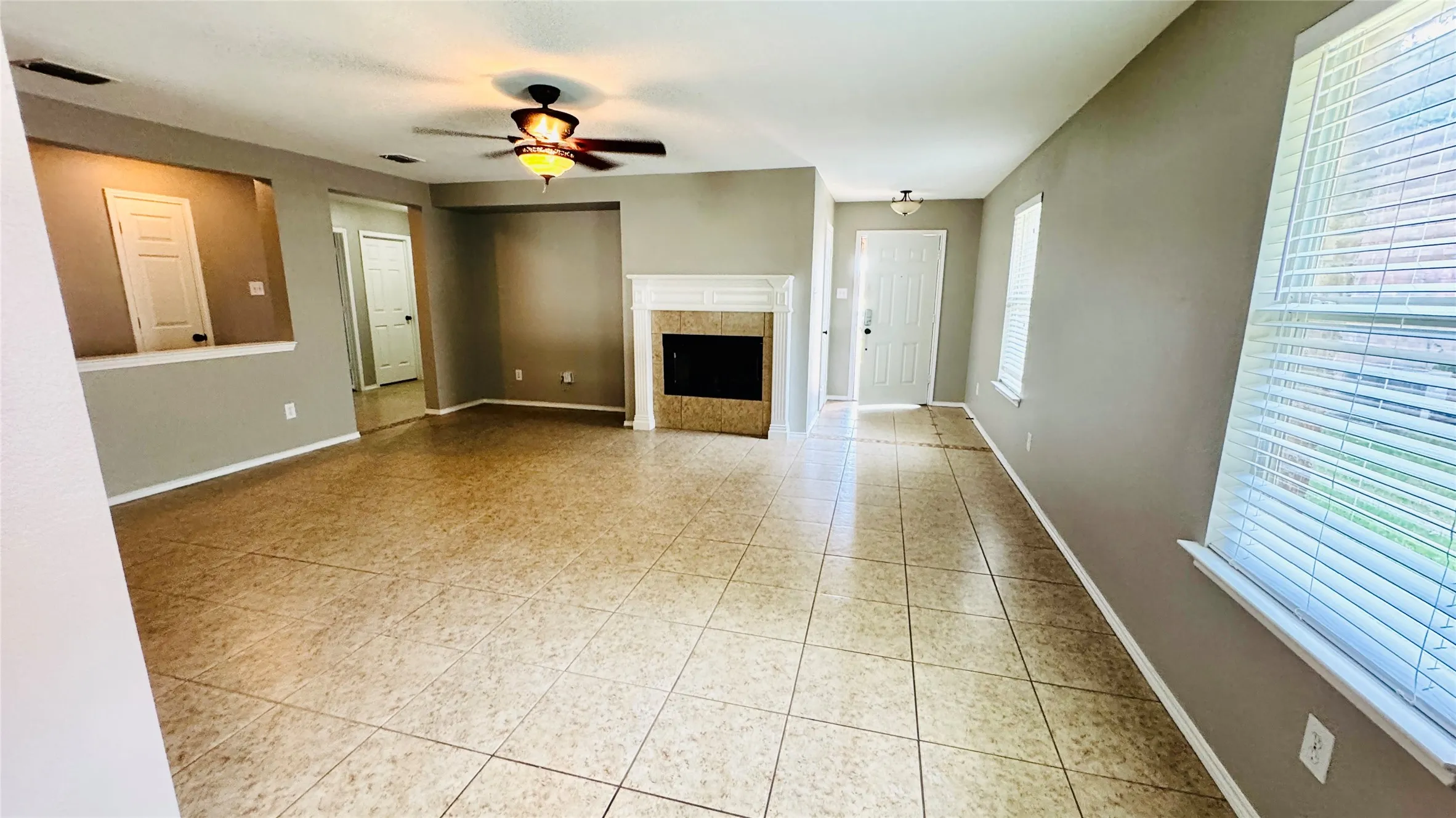 Spacious living area featuring neutral-toned tile flooring, a fireplace with a white mantel, and a ceiling fan with integrated lighting