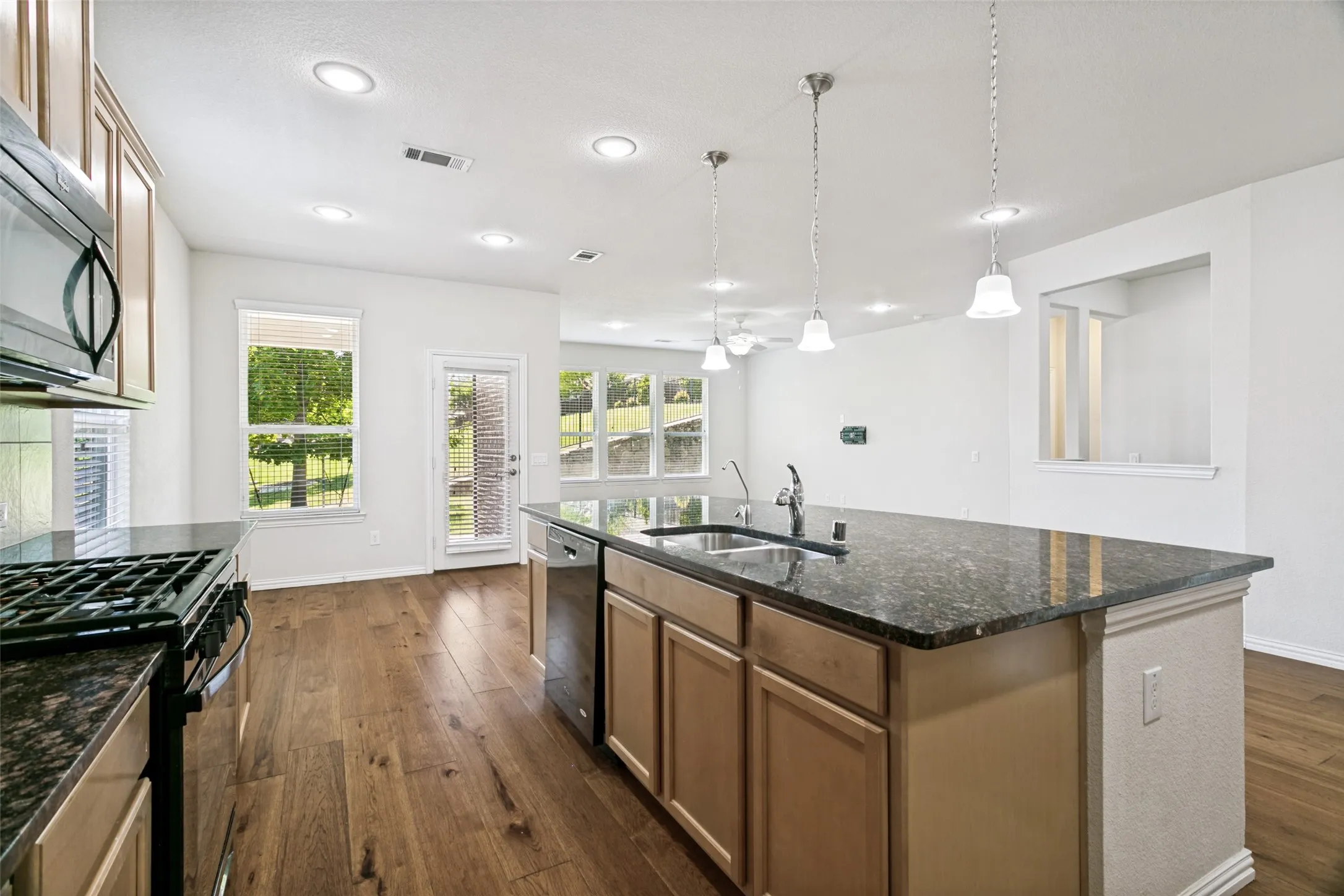 Kitchen featuring wood-finish flooring, granite countertops, light wood cabinetry, stainless steel appliances, and a central island with a double basin sink and pendant lighting
