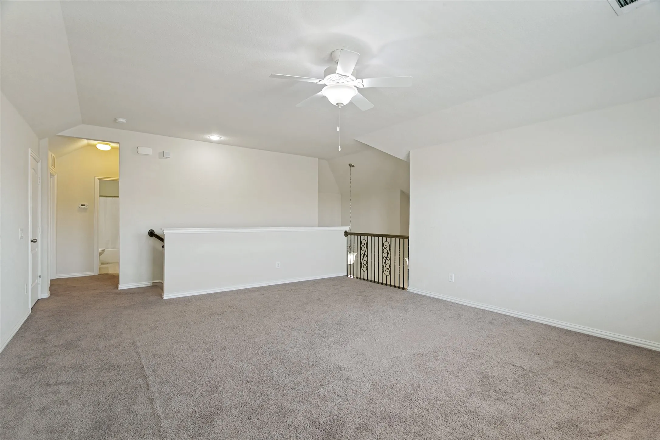 Spacious carpeted upper-level area featuring a ceiling fan with integrated lighting, a half-wall partition, and decorative metal railing