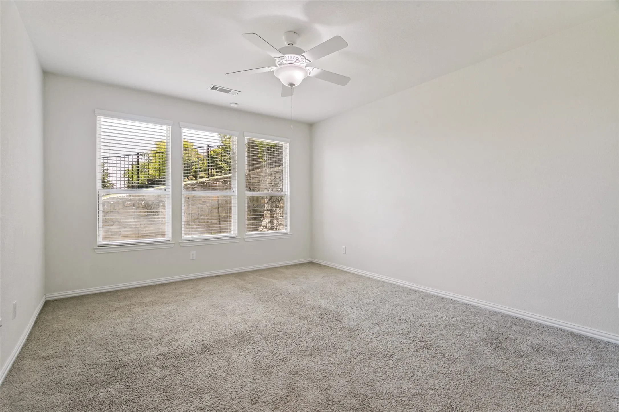 Spacious room featuring three large windows with blinds, carpeted flooring, a ceiling fan, and white baseboards