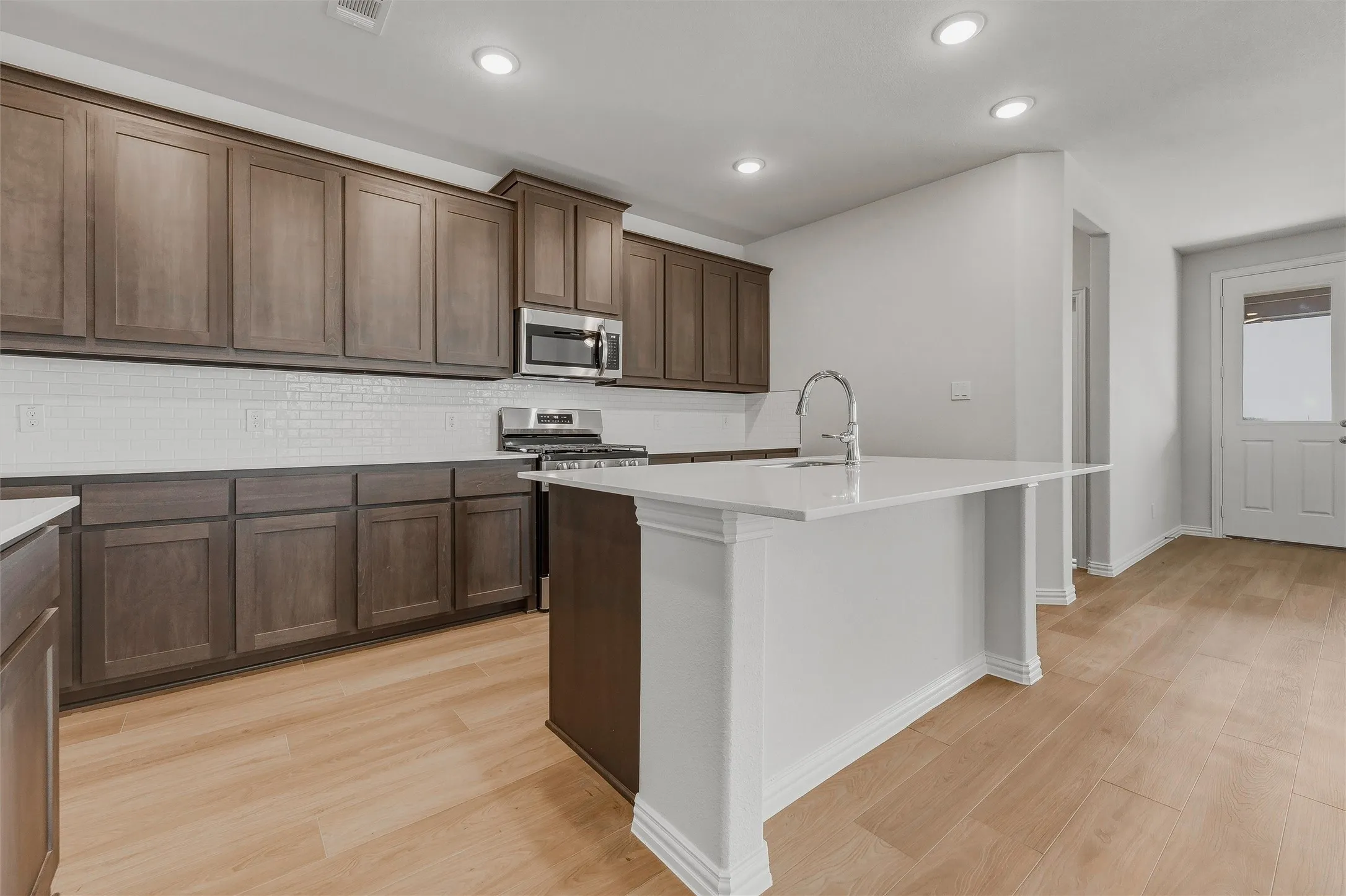 Kitchen featuring wood-finish flooring, dark wood cabinetry, white subway tile backsplash, stainless steel appliances, and a central island with a sink