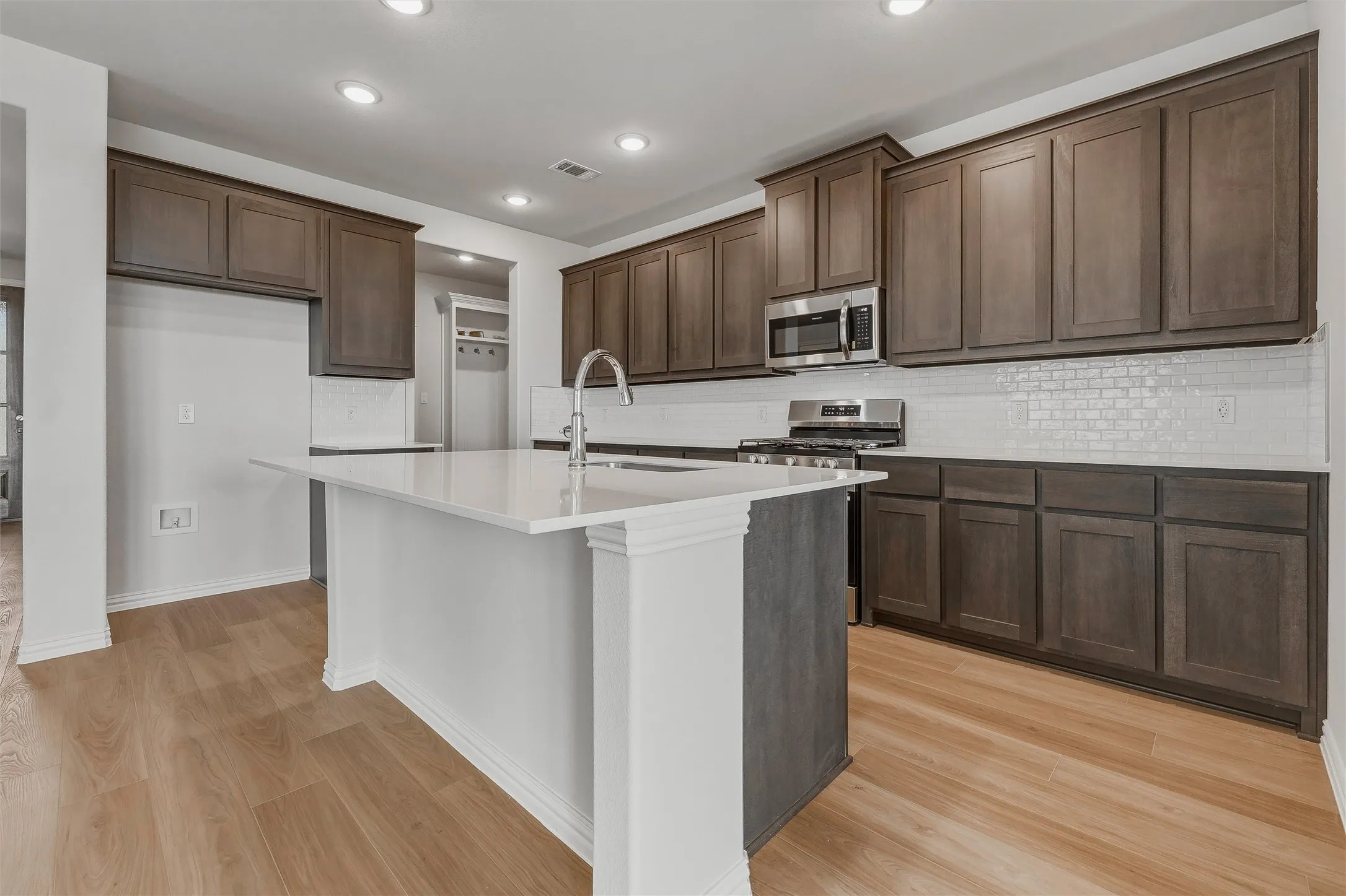 Spacious kitchen featuring an island with a sink, wood-finish flooring, white subway tile backsplash, and dark wood cabinetry