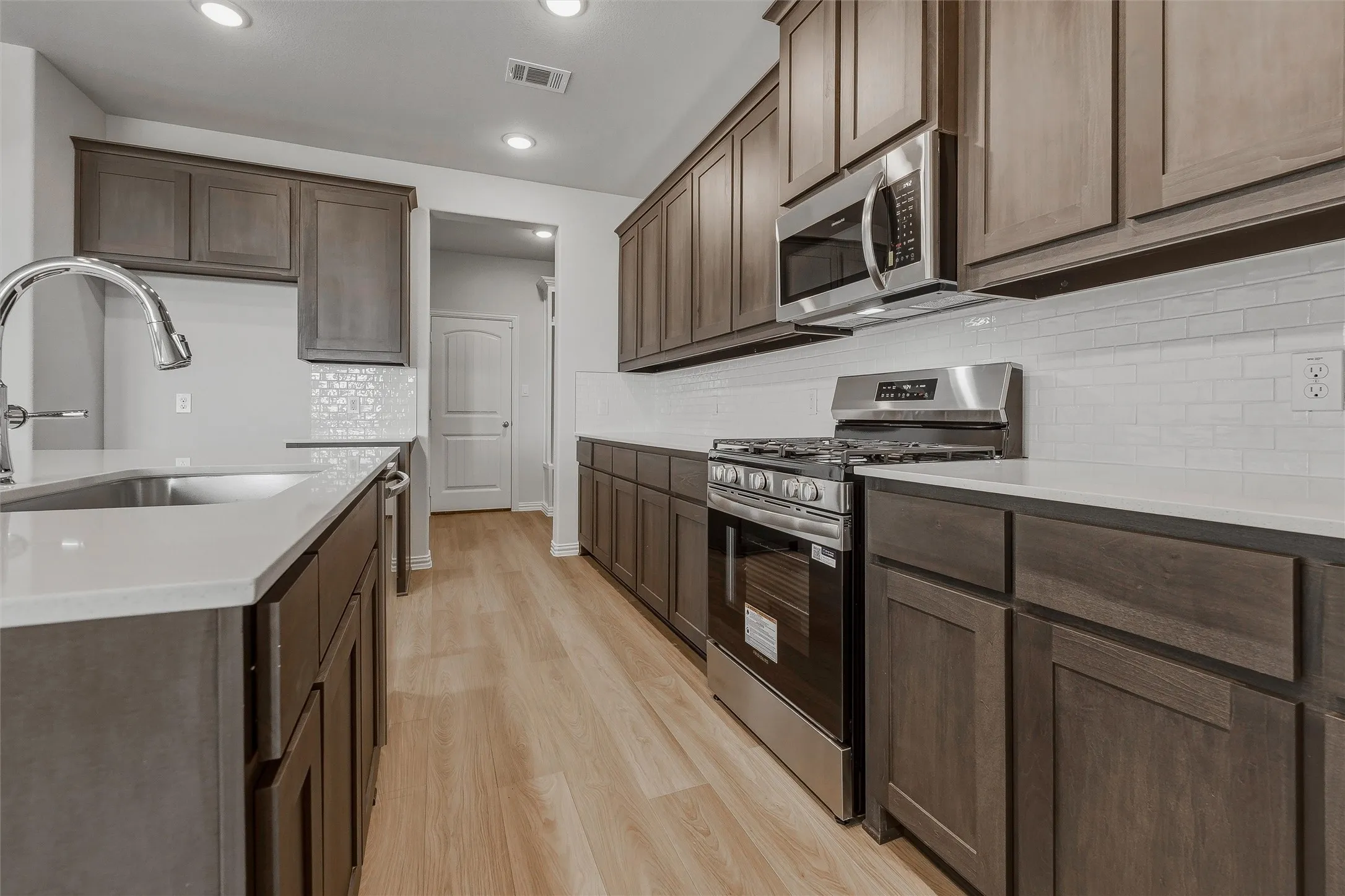 Updated kitchen featuring wood-finish cabinetry, stainless steel appliances, white subway tile backsplash, light-toned countertops, and wood-finish flooring