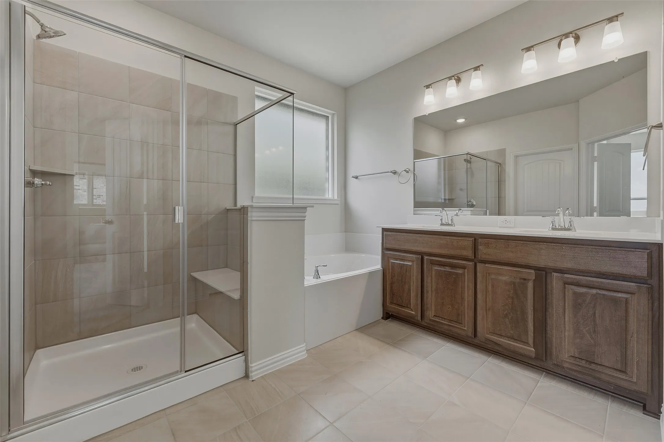 Spacious bathroom featuring a glass-enclosed shower with integrated bench, a separate soaking tub, a double vanity with ample storage, and large format floor tiling