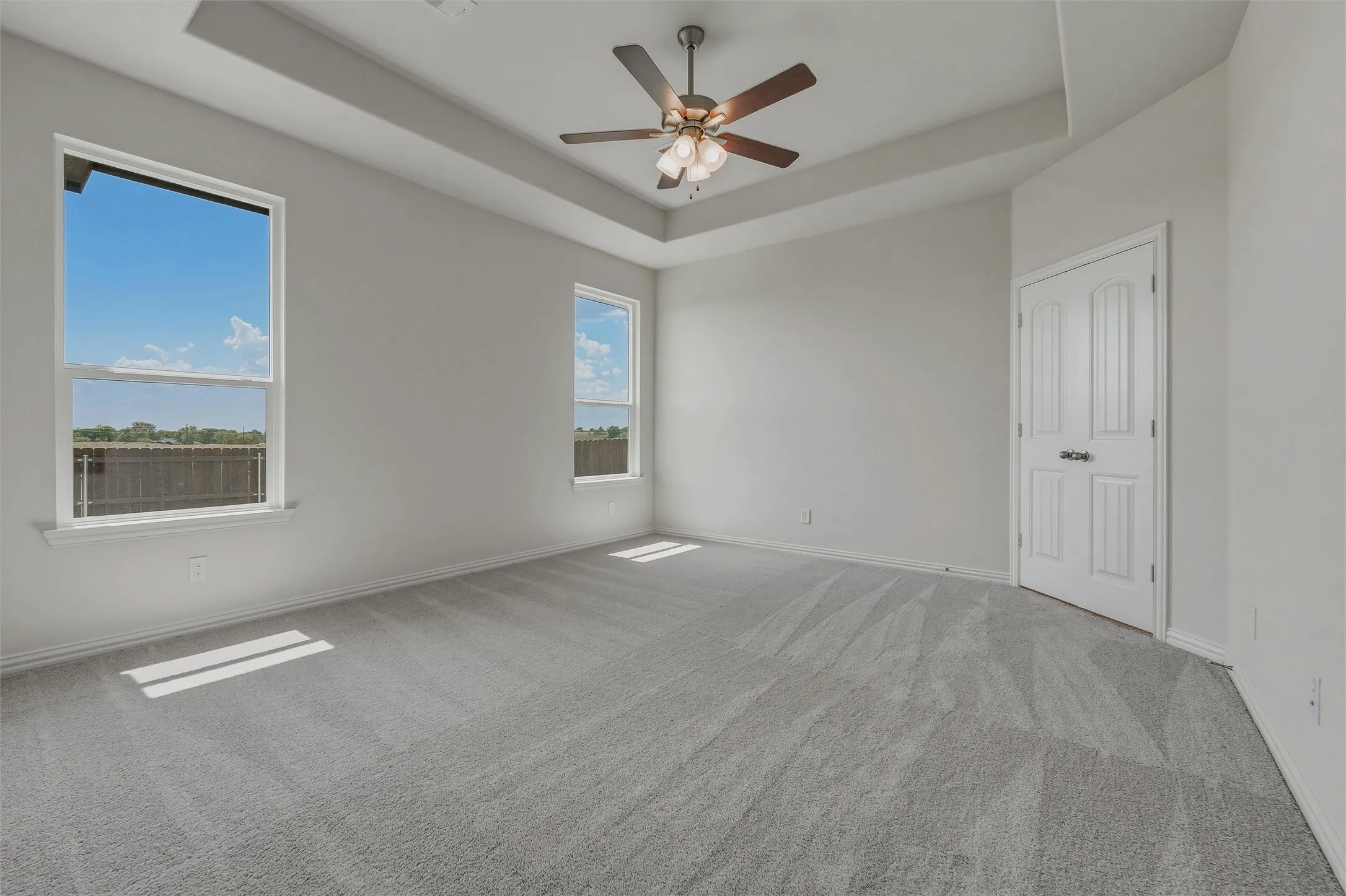 Recessed tray ceiling, ceiling fan with light fixture, gray carpet, white double-pane windows, and a paneled door