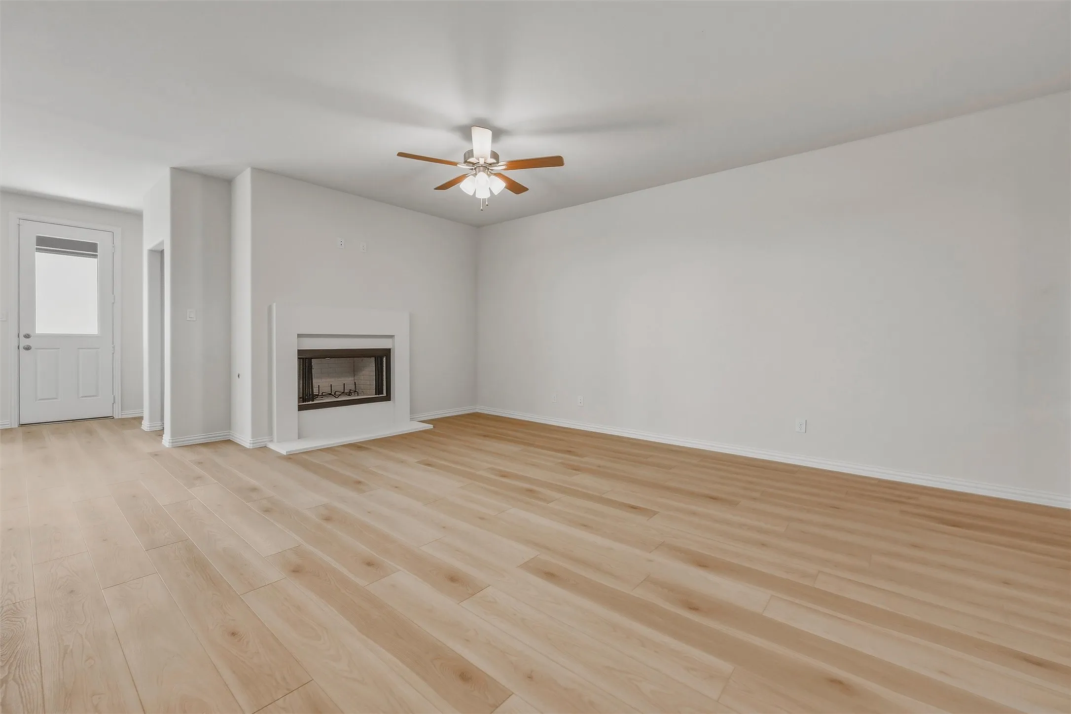 Light wood-finish flooring, a fireplace with a hearth, a ceiling fan with light fixture, and a white paneled door with glass insert