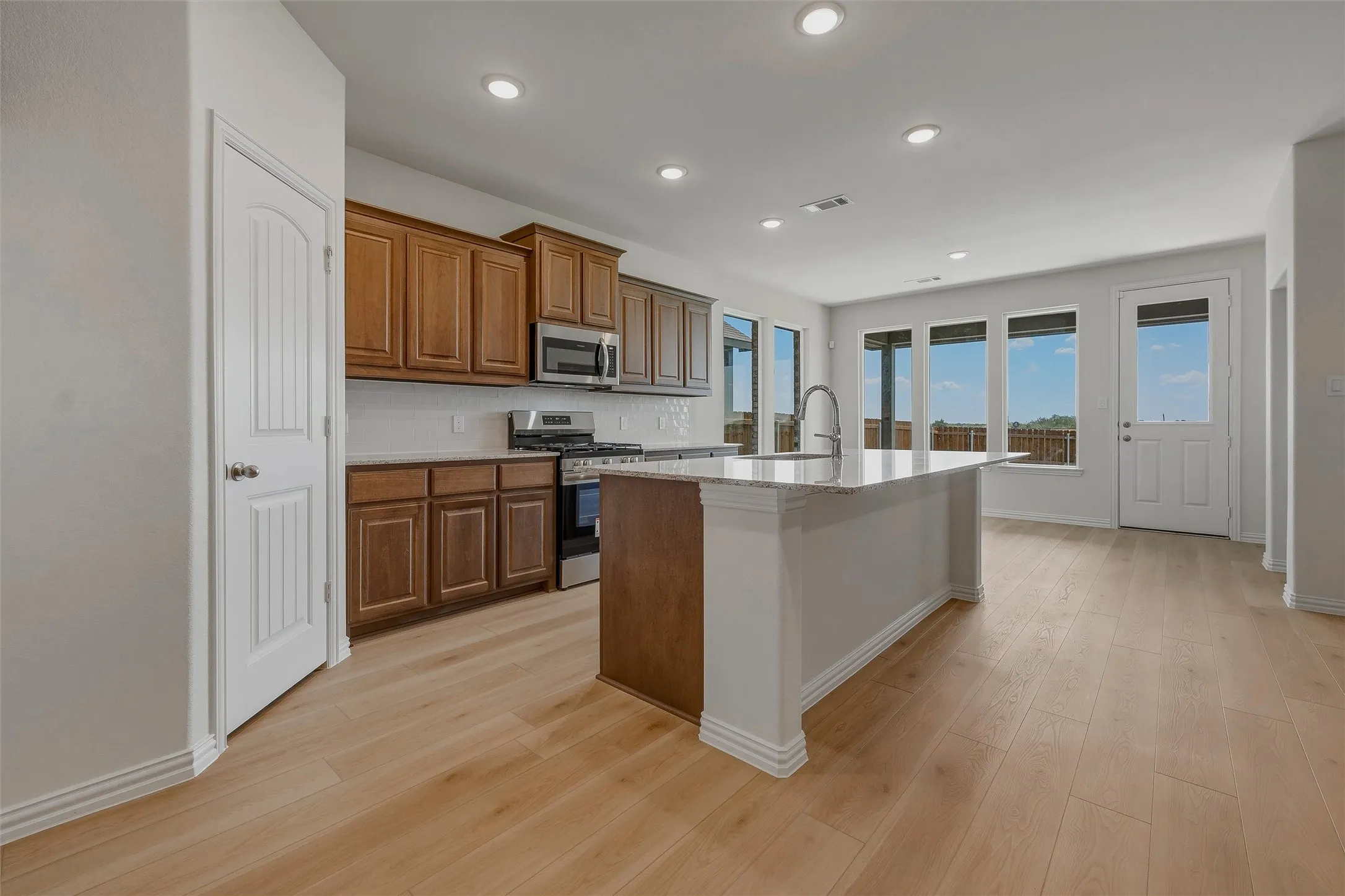 Light wood-finish flooring throughout, recessed ceiling lighting, kitchen island with built-in sink, stainless steel appliances, and wood cabinetry
