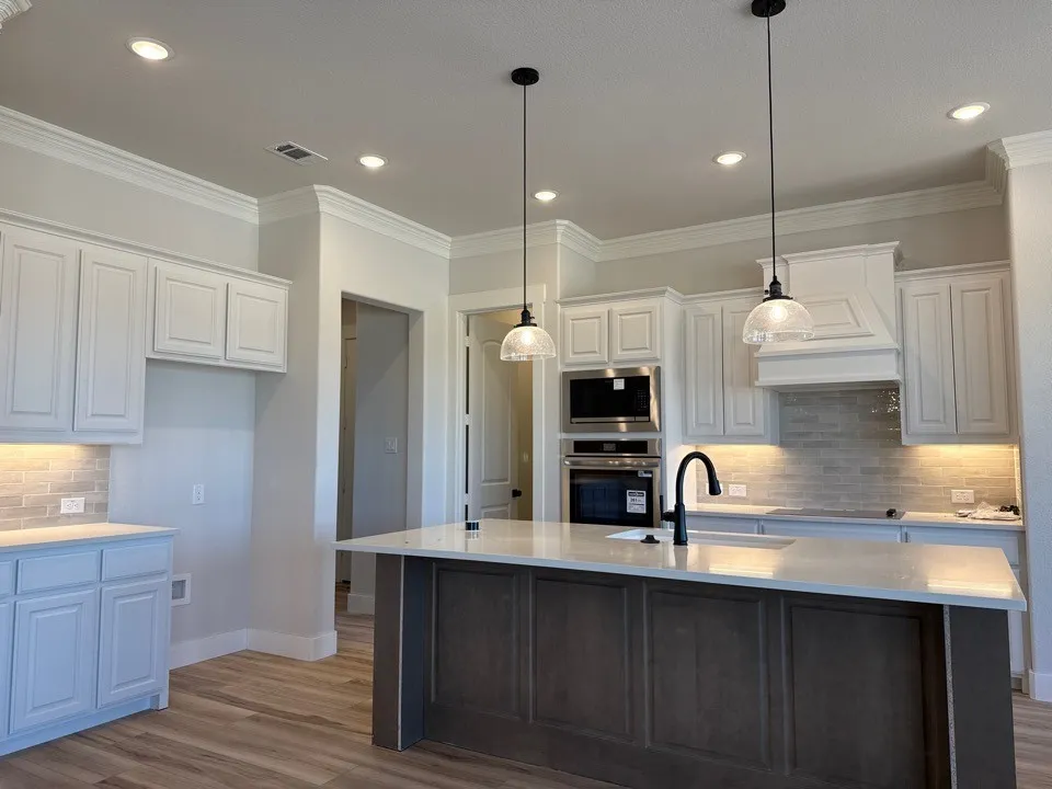 White shaker-style cabinetry, wood-finish flooring, built-in stainless steel appliances, a center island with a sink, and pendant lighting