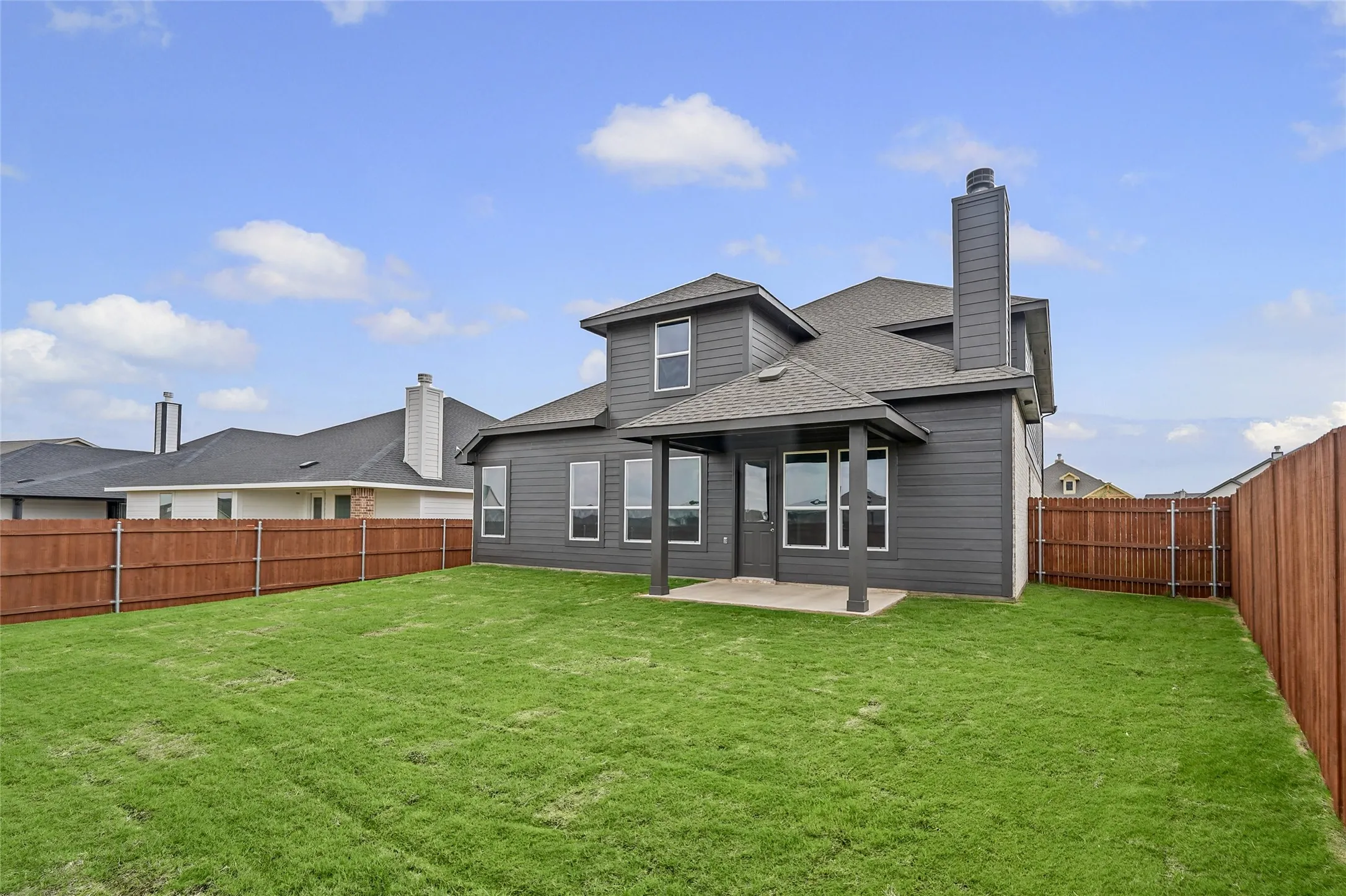 Expansive backyard featuring a manicured lawn, wood privacy fencing, a covered patio, and a two-story exterior with a prominent chimney