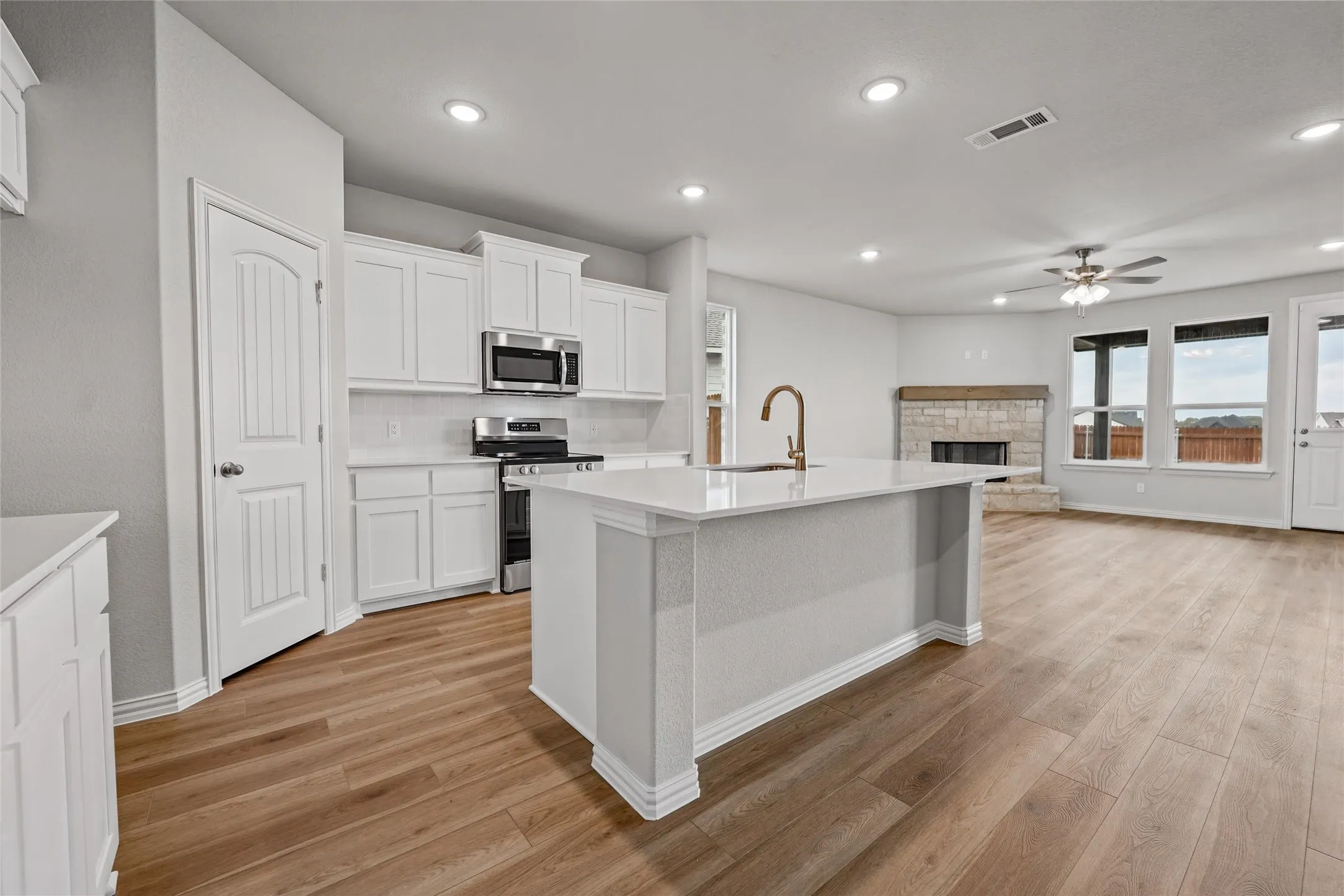 Open-concept living space featuring wood-finish flooring, a kitchen island with a single basin sink, white cabinetry with stainless steel appliances, and a stone-clad fireplace