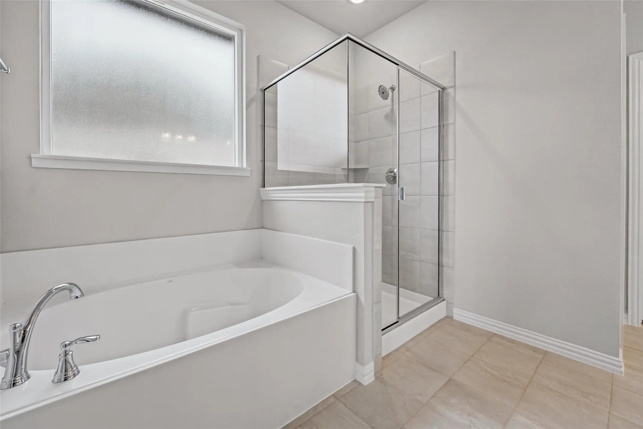 Built-in soaking tub with chrome fixtures, enclosed glass shower with tiled walls, frosted window, and neutral tile flooring