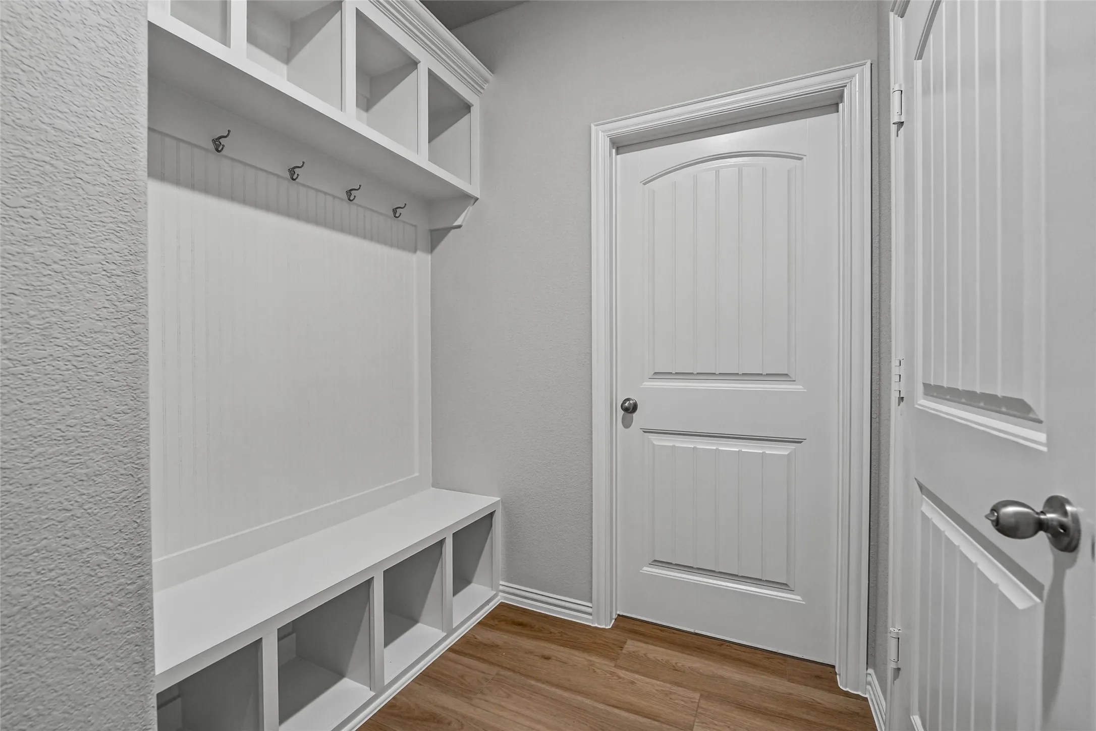 Built-in storage bench with cubbies, overhead shelving, wall-mounted hooks, beadboard paneling, and wood-finish flooring
