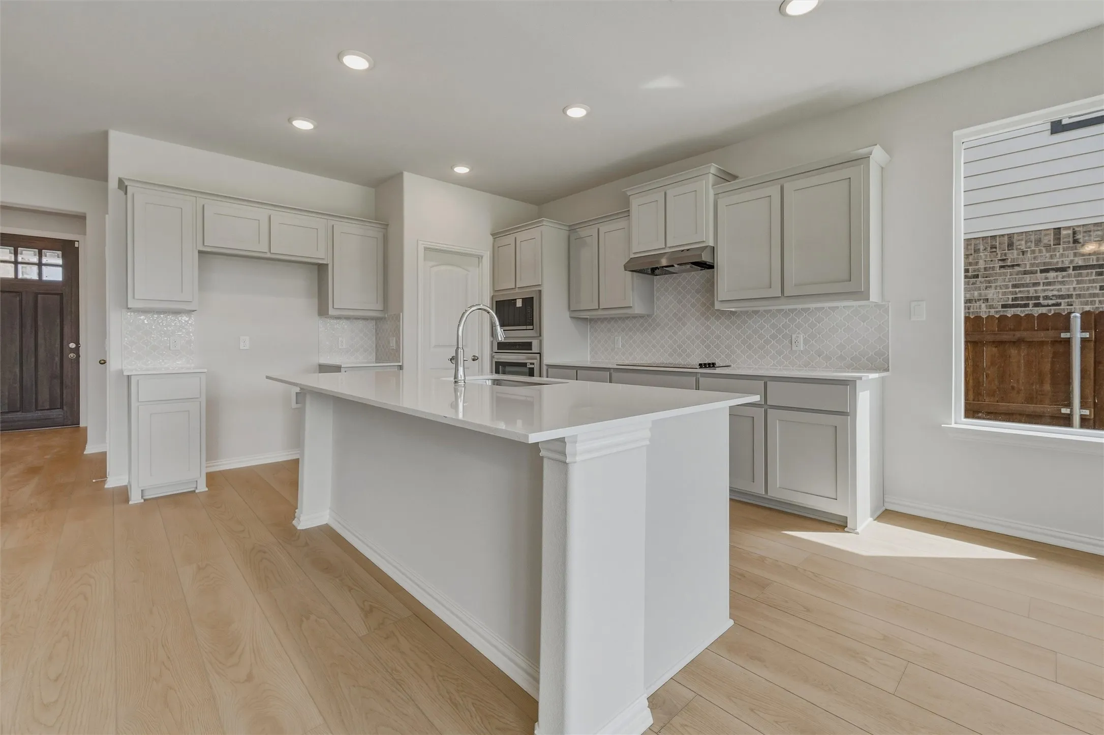 Spacious kitchen featuring an expansive central island, light cabinetry, a patterned tile backsplash, stainless steel appliances, and light wood-finish flooring