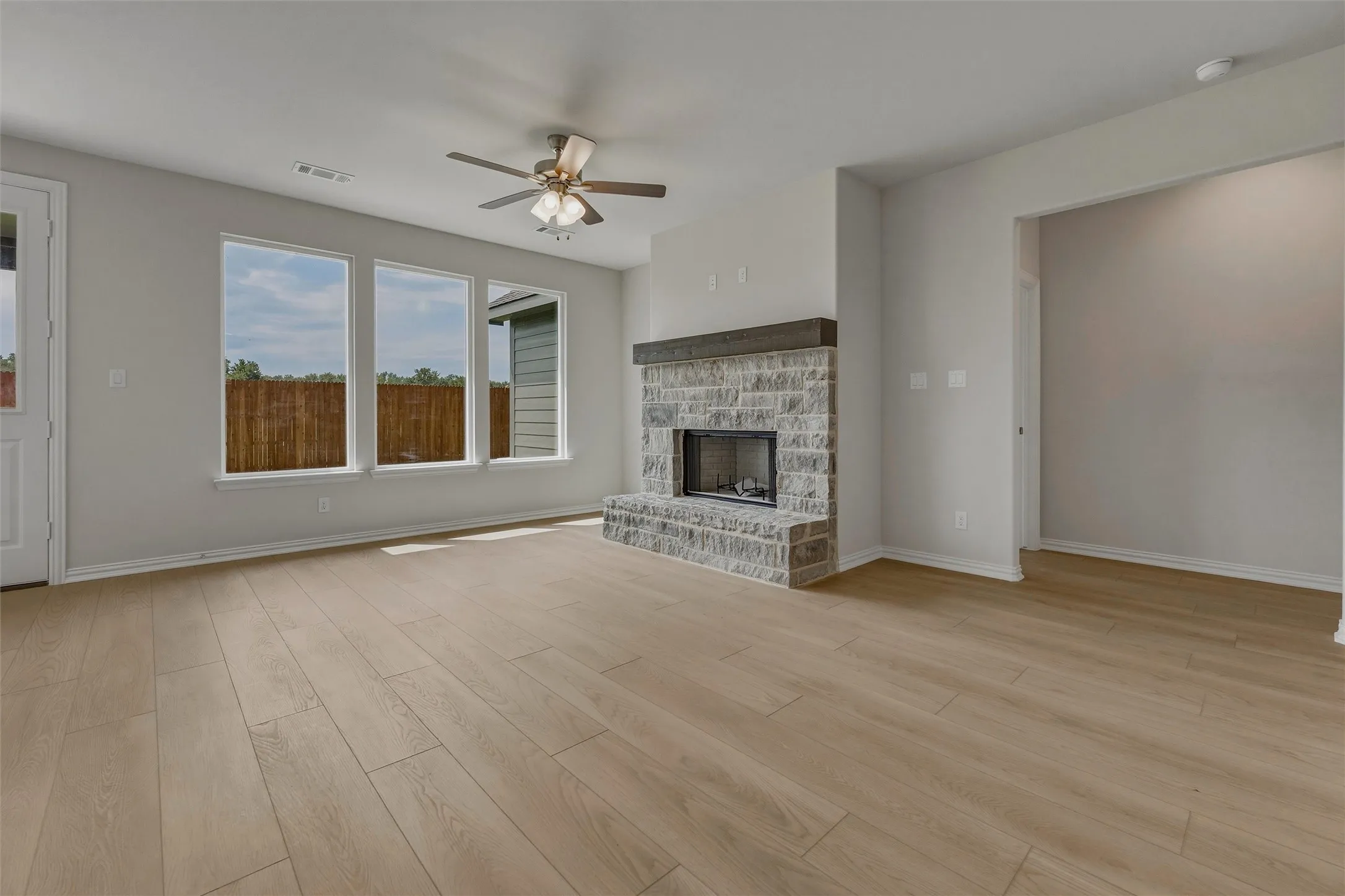 Spacious living area featuring light wood-finish flooring, a stone-clad fireplace with a wood mantle, a ceiling fan, and multiple windows